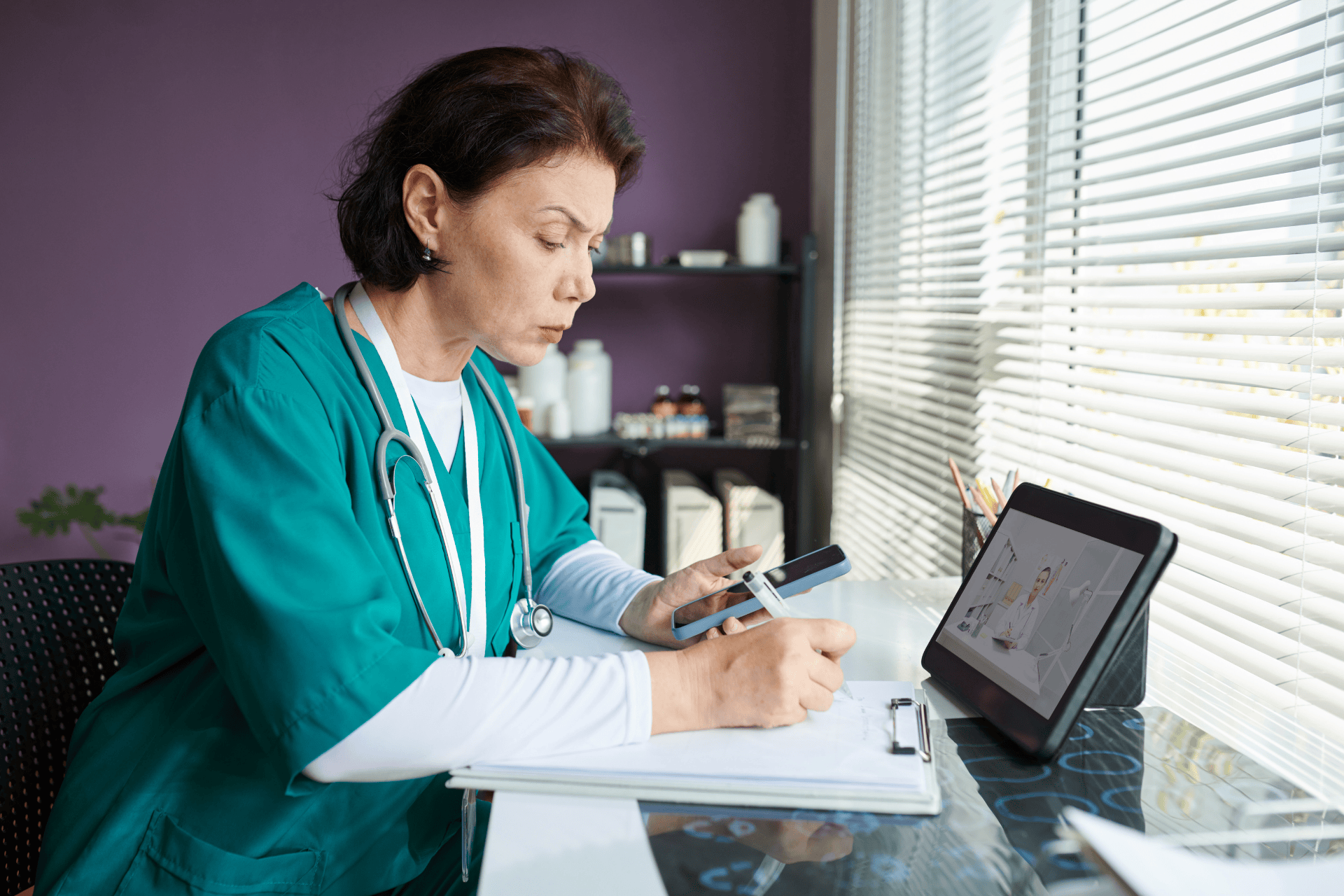 Female doctor in scrubs reviewing a phone while taking notes during a telehealth session.