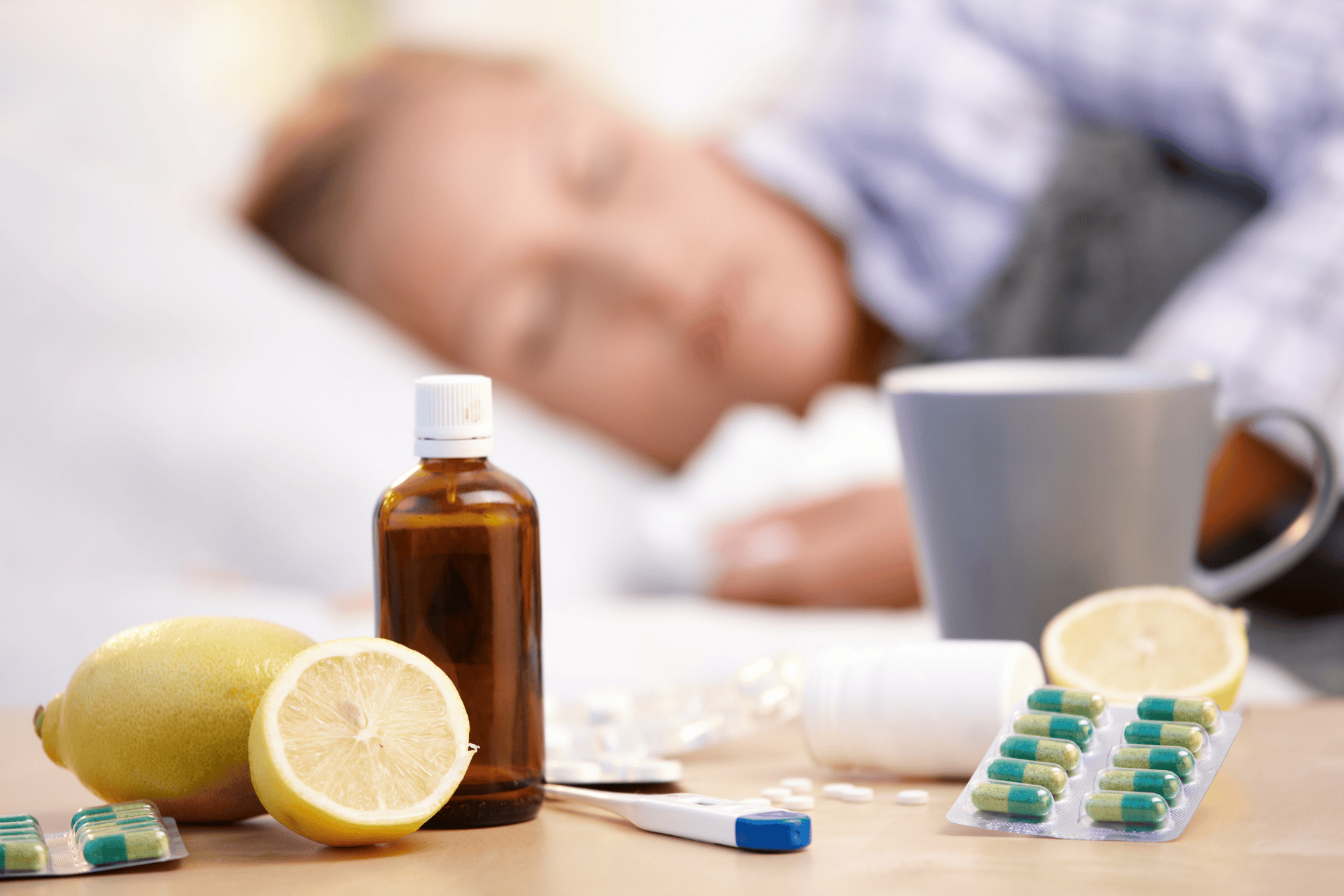 Medicine, lemon, thermometer, and pills on a bedside table with a sick person resting in the background.