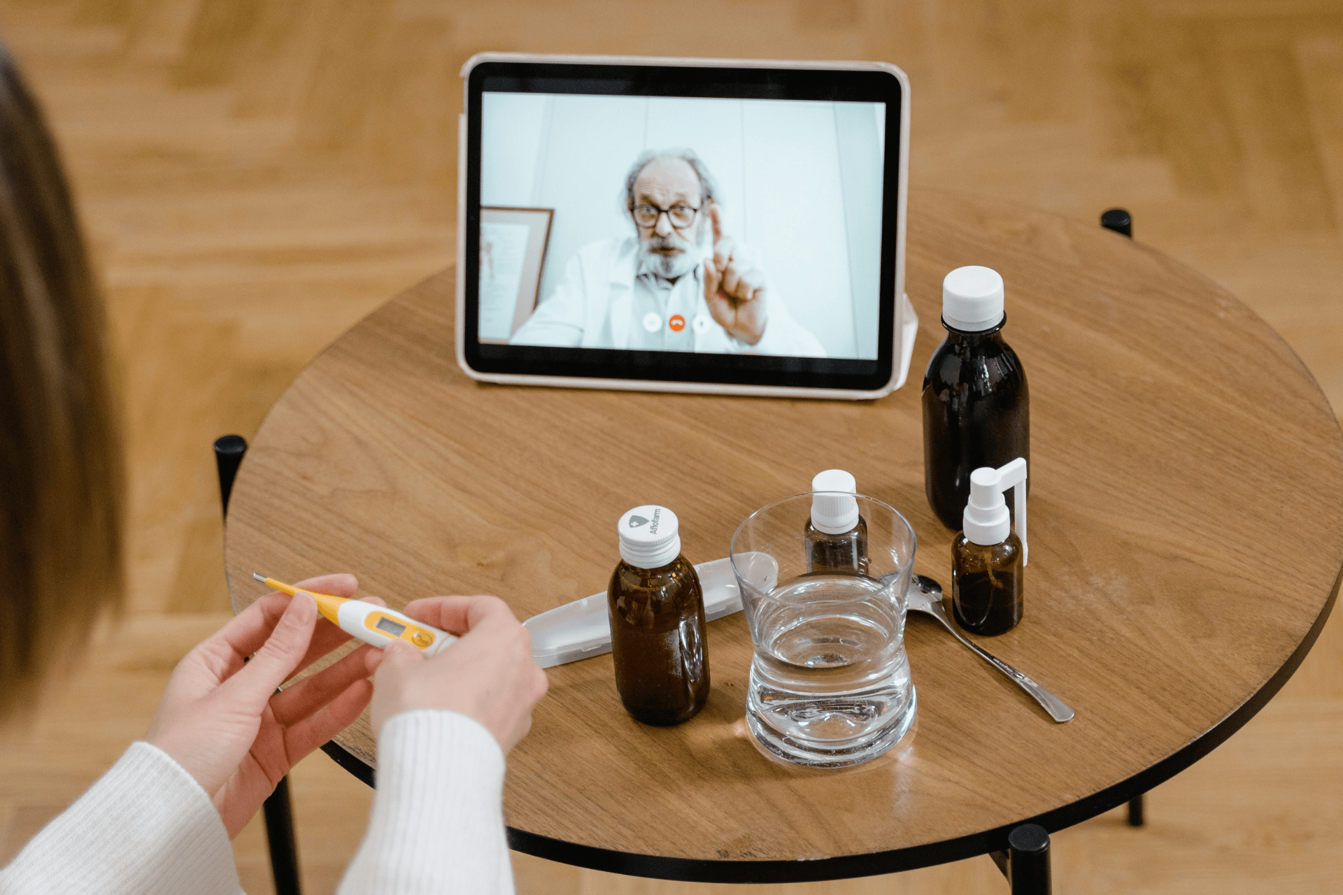 Person holding a thermometer while having an online consultation with a doctor on a tablet, with medicine bottles and a glass of water on the table.