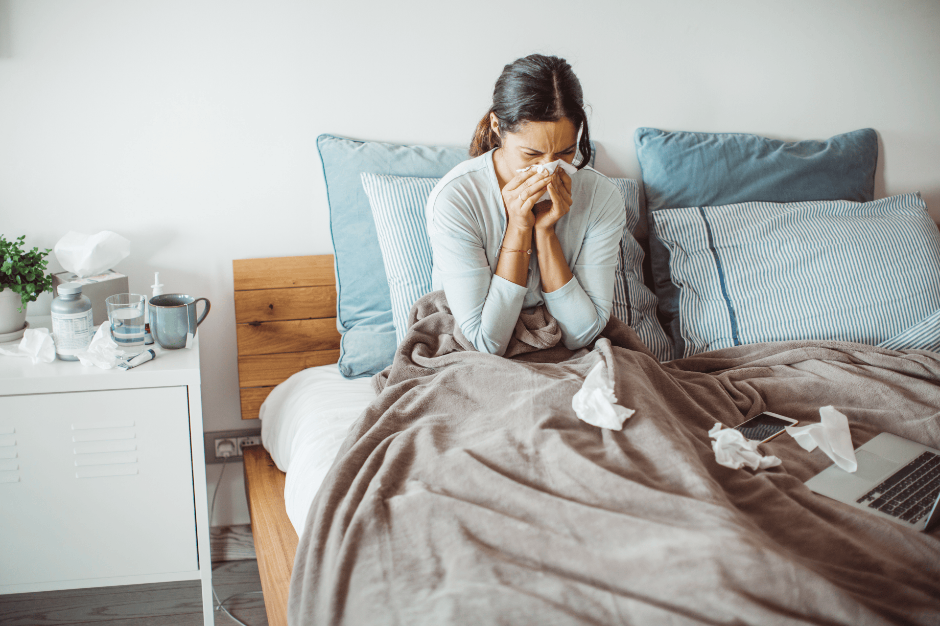 Woman sitting in bed blowing her nose, surrounded by tissues and medicine.