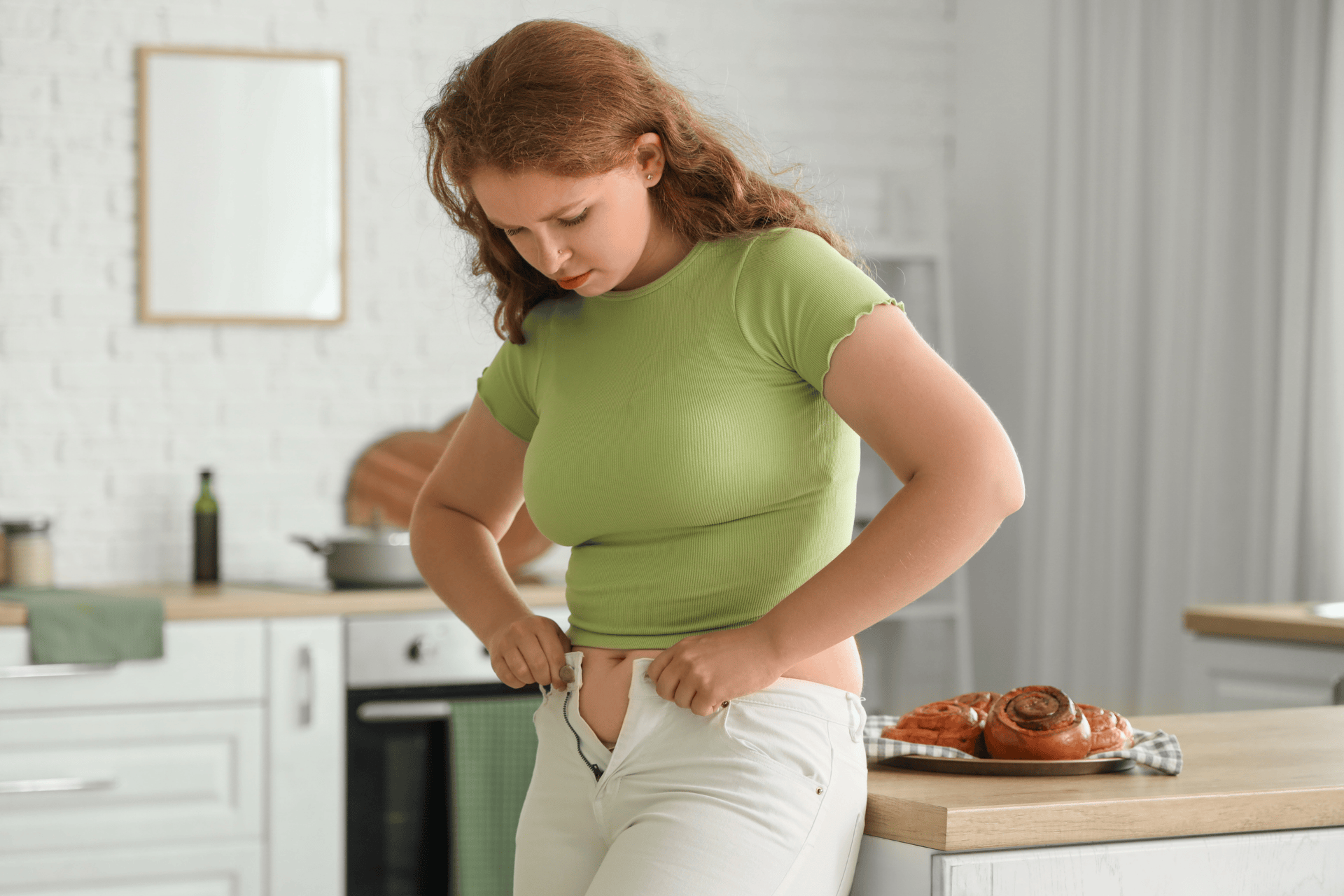 Woman struggling to button tight pants in a kitchen.