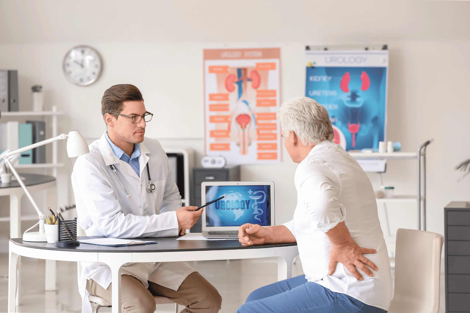 Doctor showing a urology diagram on a laptop to an older male patient holding his lower back in a medical office.