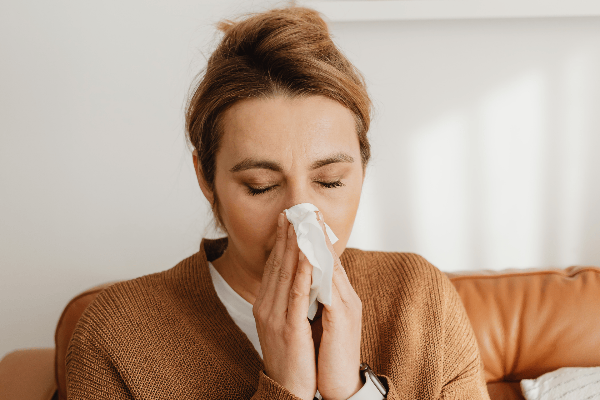 Woman blowing her nose into a tissue while sitting on a couch.