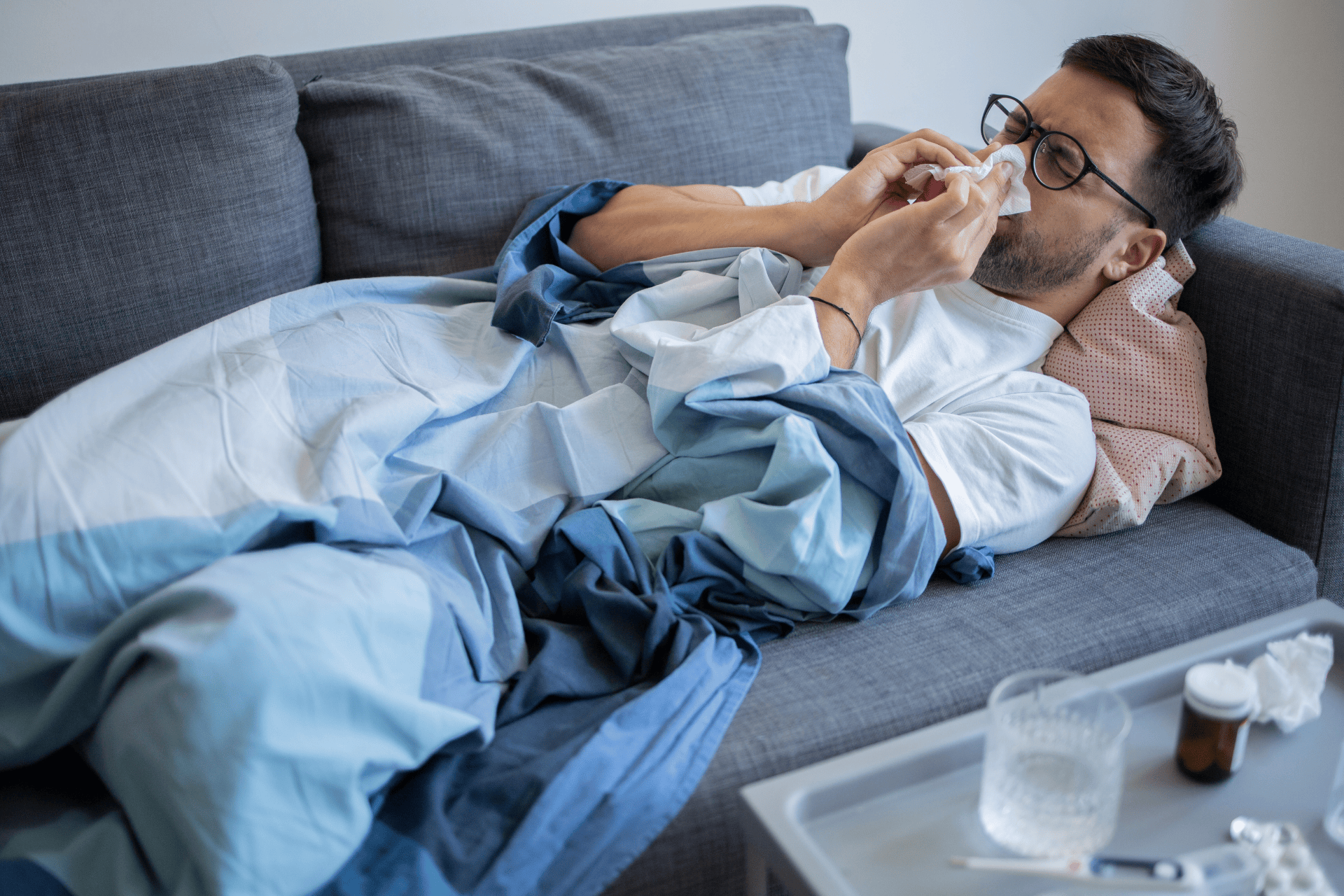 Man lying on a couch under a blanket, blowing his nose, with cold medicine and tissues on a nearby tray.