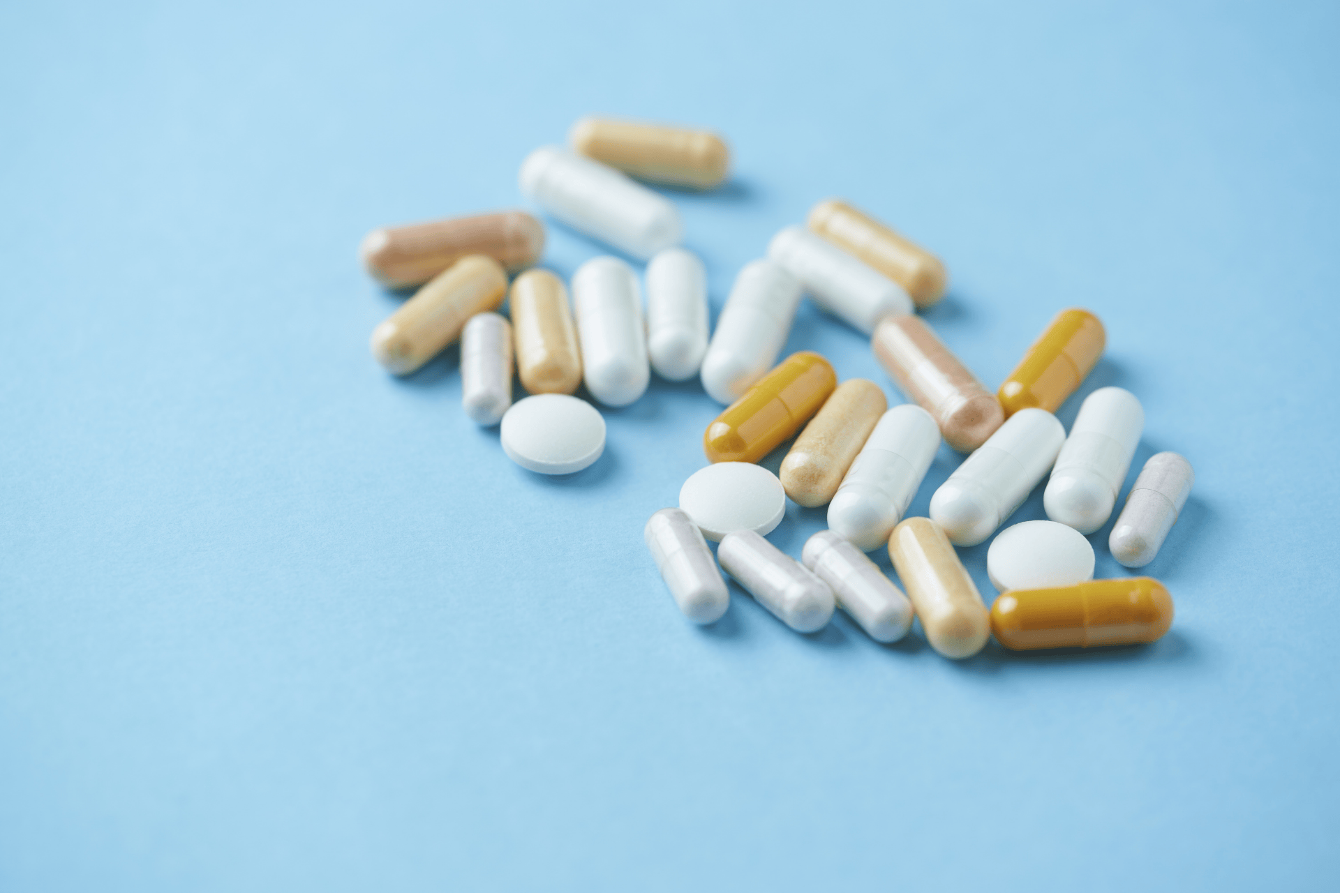 A scattered assortment of capsules and tablets in various colors—white, beige, gold, and light brown lying on a light blue surface.