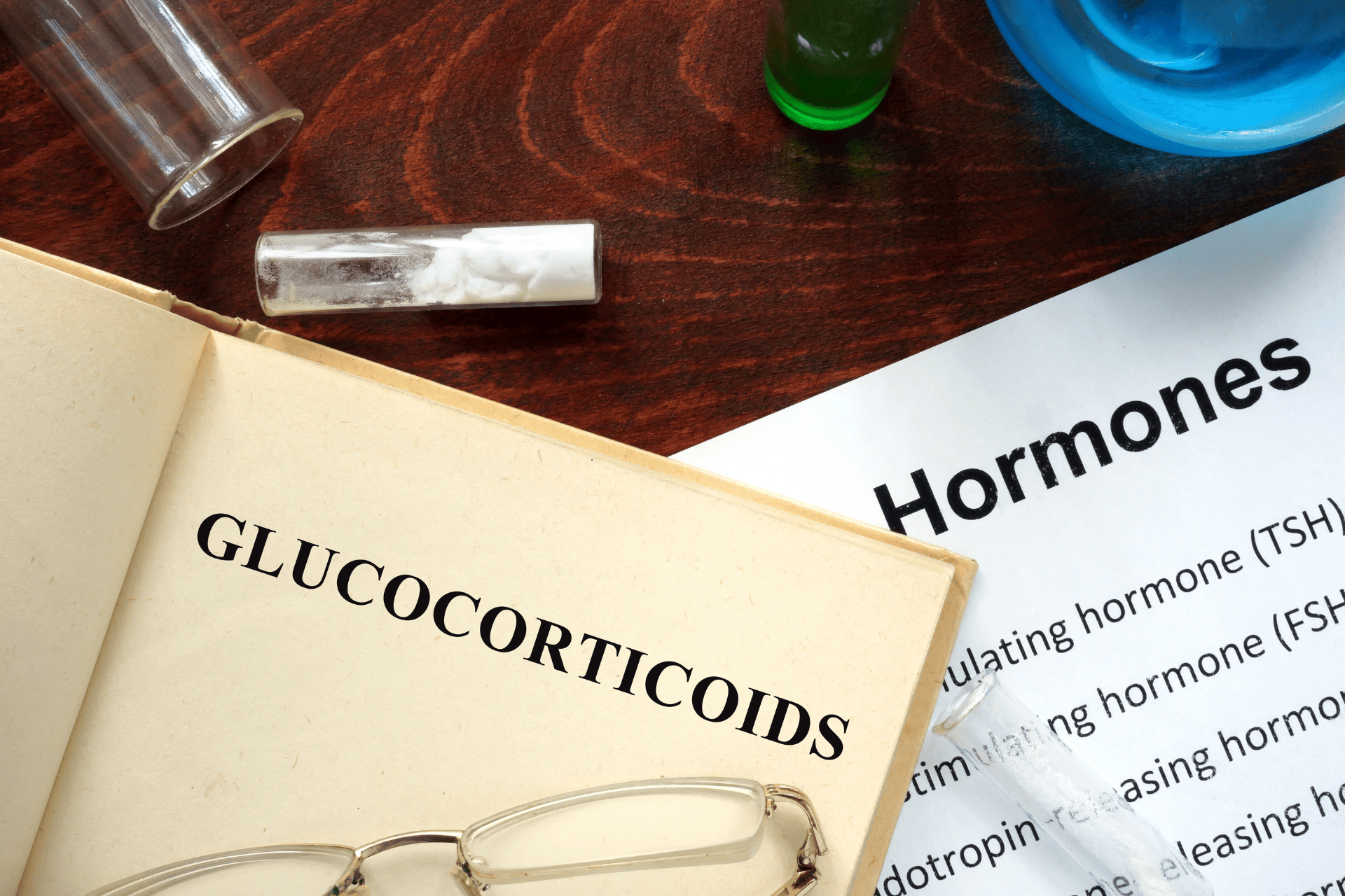 Open book labeled “GLUCOCORTICOIDS” next to a “Hormones” sheet and lab vials on a table.