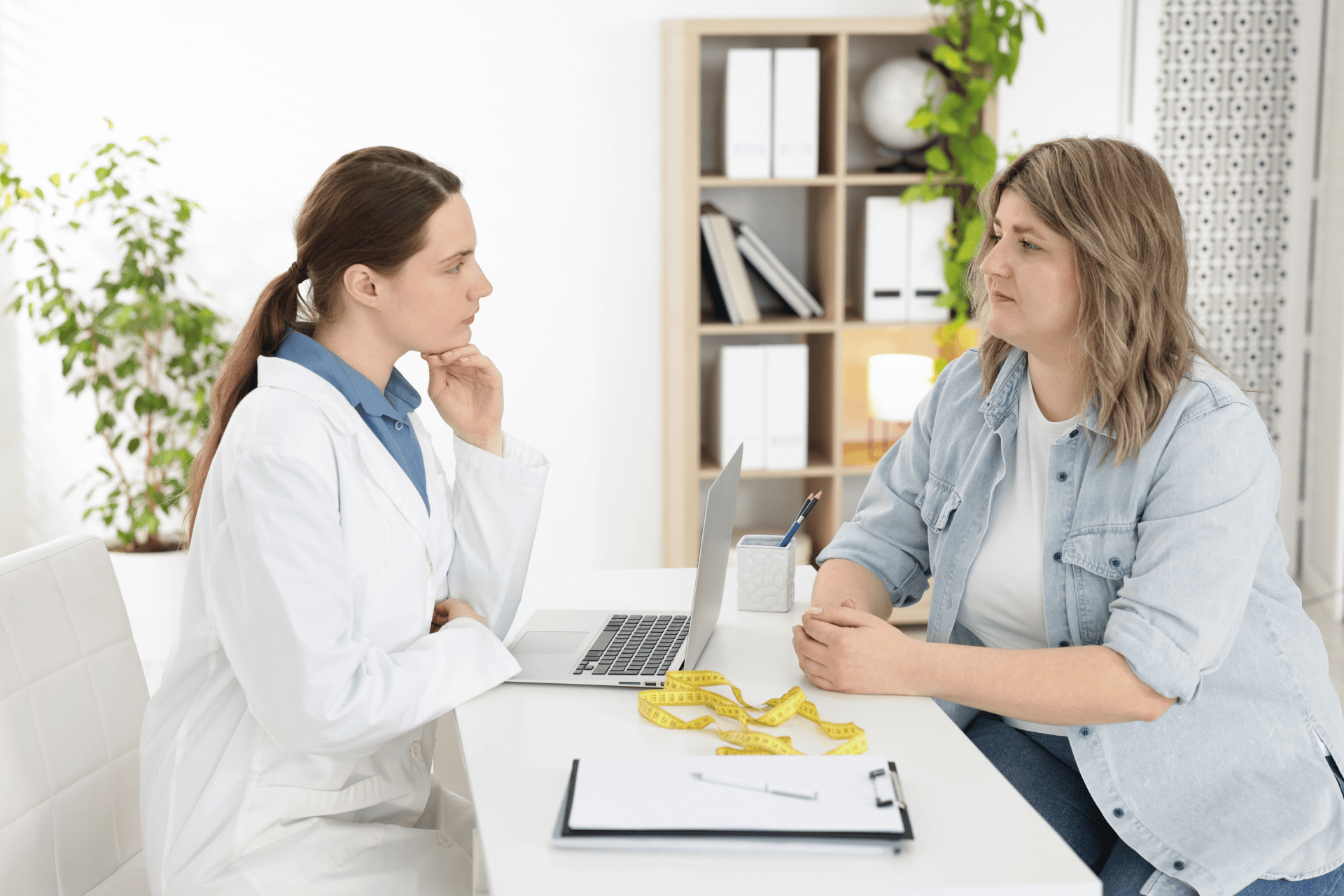 A doctor sits across from a patient at a desk, discussing health concerns; a laptop, a clipboard, and a measuring tape are on the table.