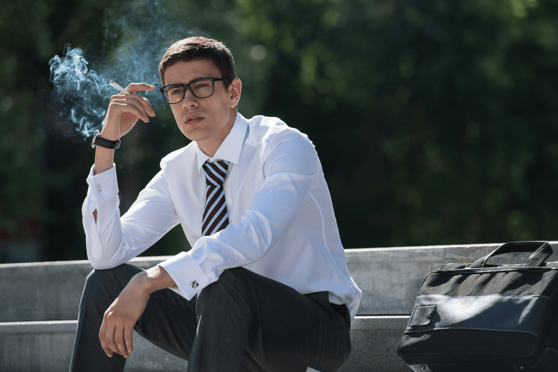 A man in business attire, wearing glasses and sitting outdoors on steps, holds a lit cigarette with smoke rising, with a briefcase beside him.
