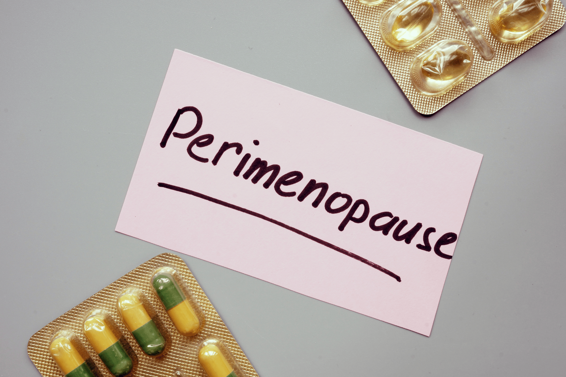 A pink card with the word "Perimenopause" handwritten on it, placed between blister packs of capsules and softgel supplements.