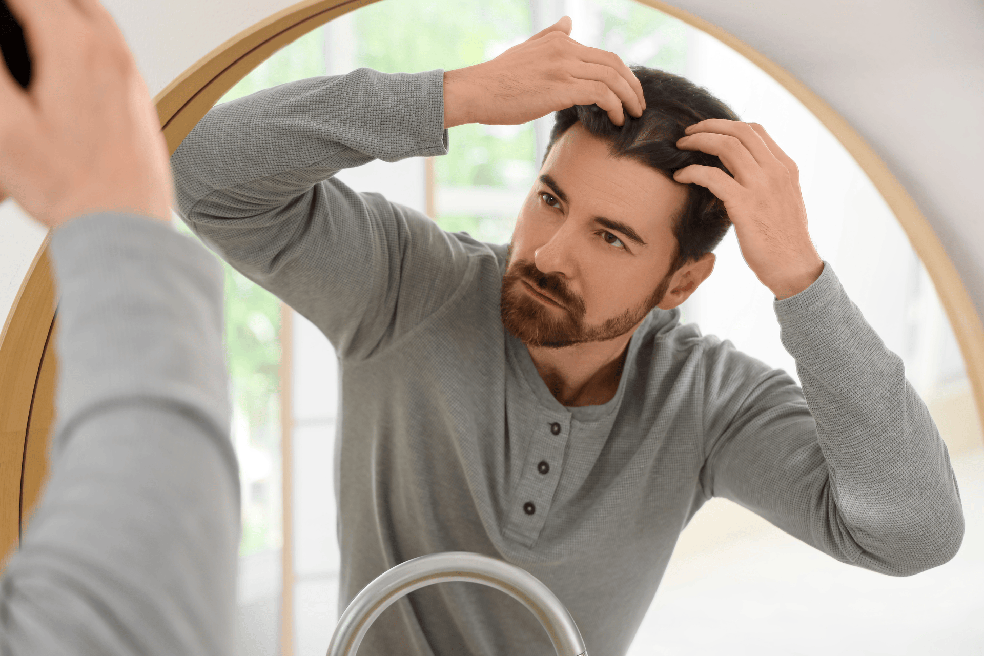 Man looking in a mirror and examining his hairline with his hands.