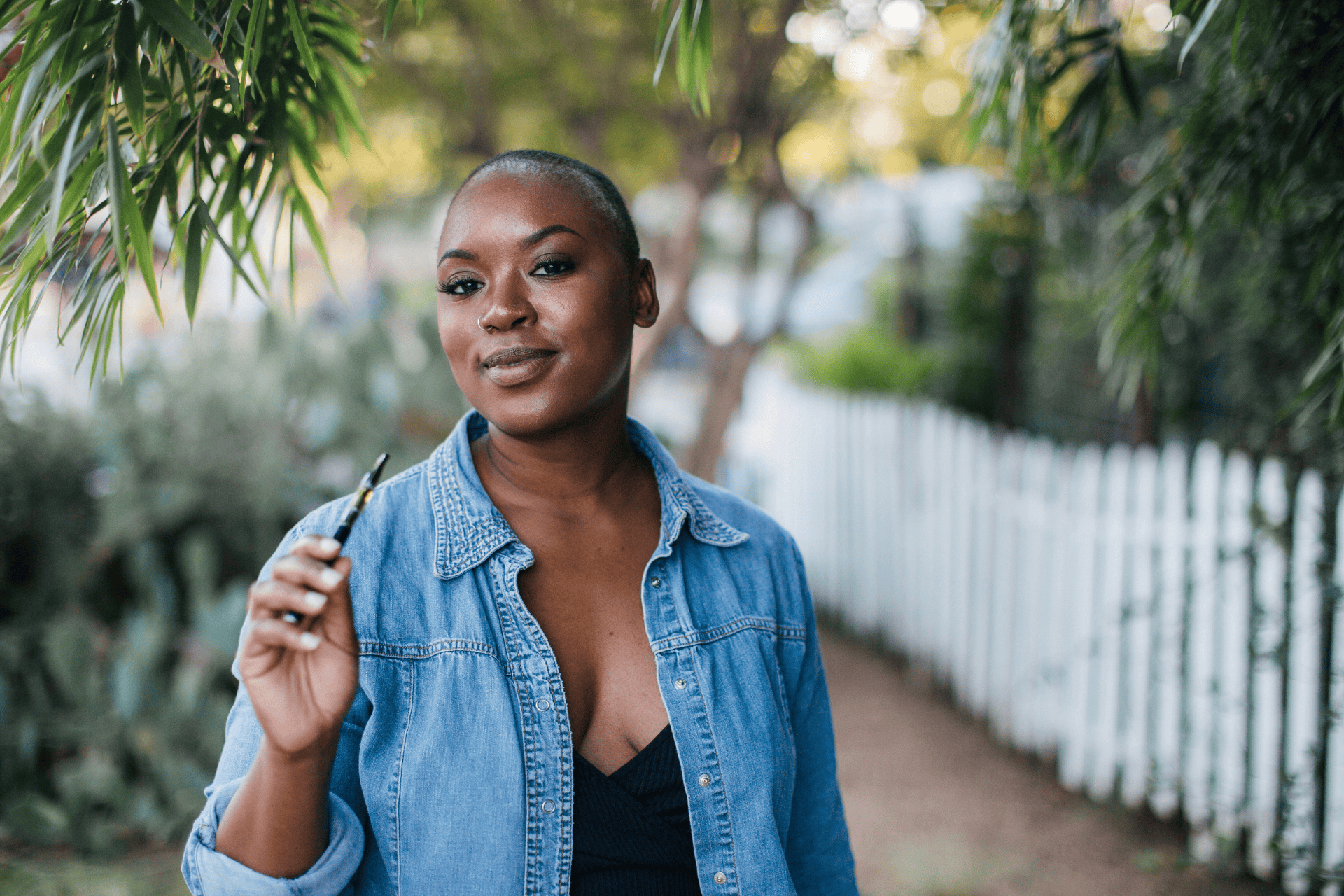 A woman standing outdoors holding a vape pen near her face.