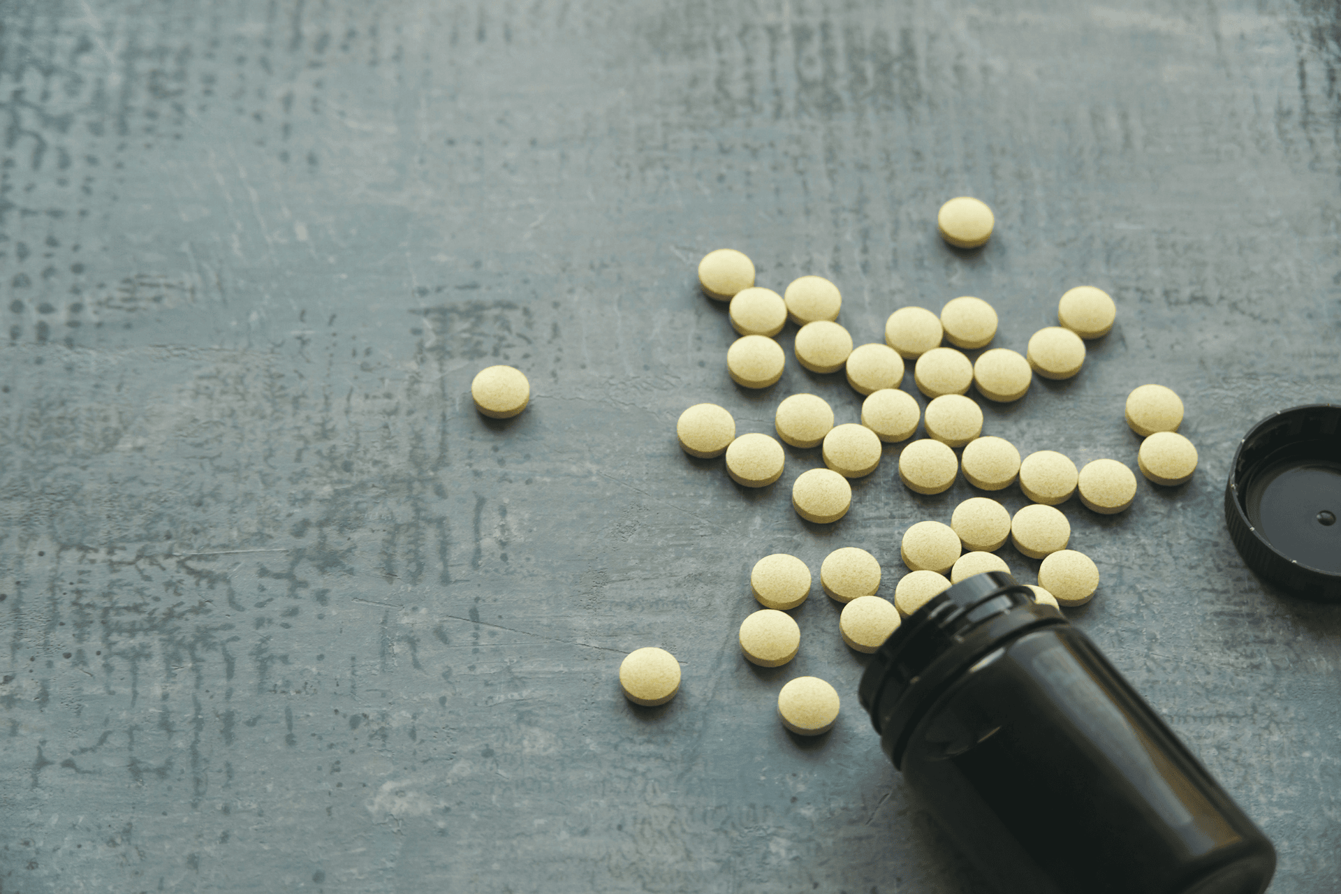 Yellow tablets spilled from a black pill bottle onto a gray surface.