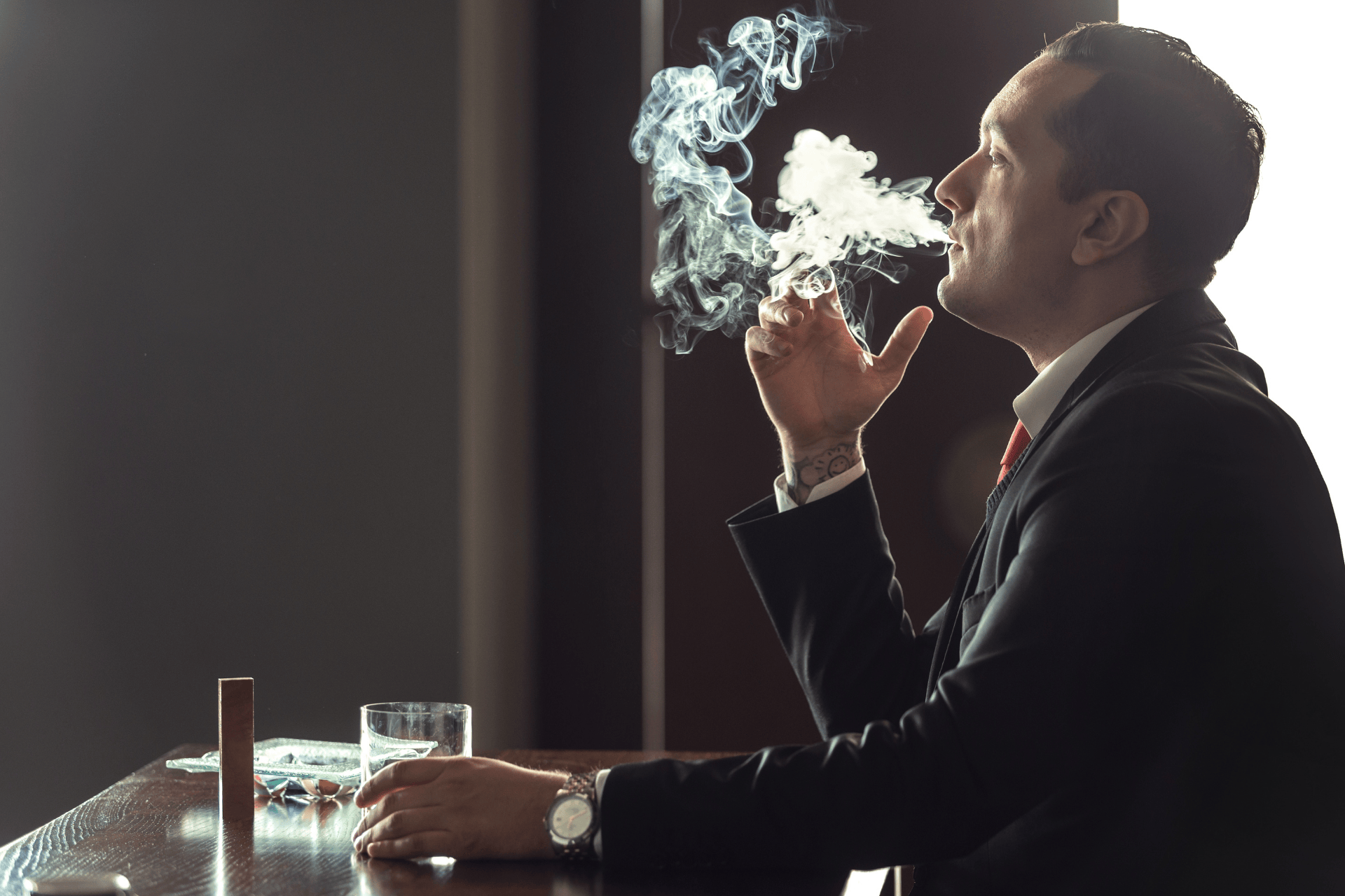 A man in a suit sitting at a table, smoking a cigarette with smoke rising.