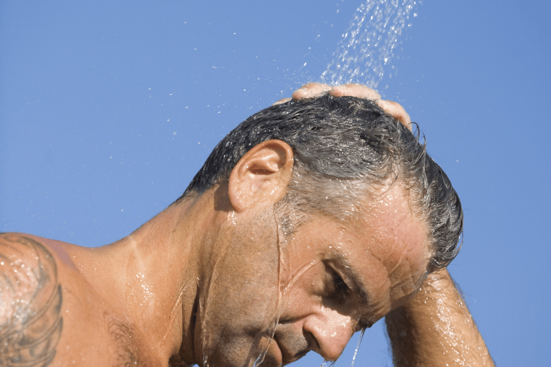 A man rinsing his hair under running water against a clear blue sky.