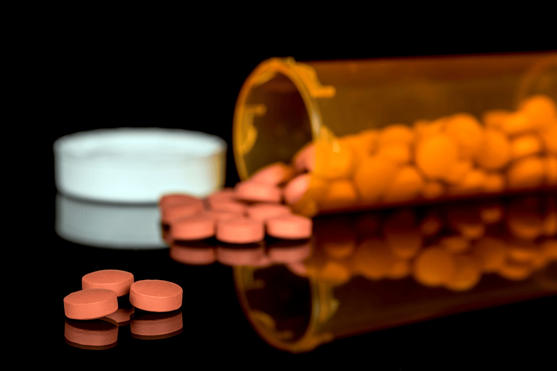 Orange prescription pills spilled from an amber pill bottle on a dark surface.