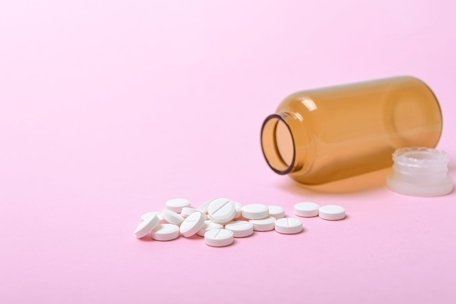 White tablets spilled from an amber pill bottle on a pink background.
