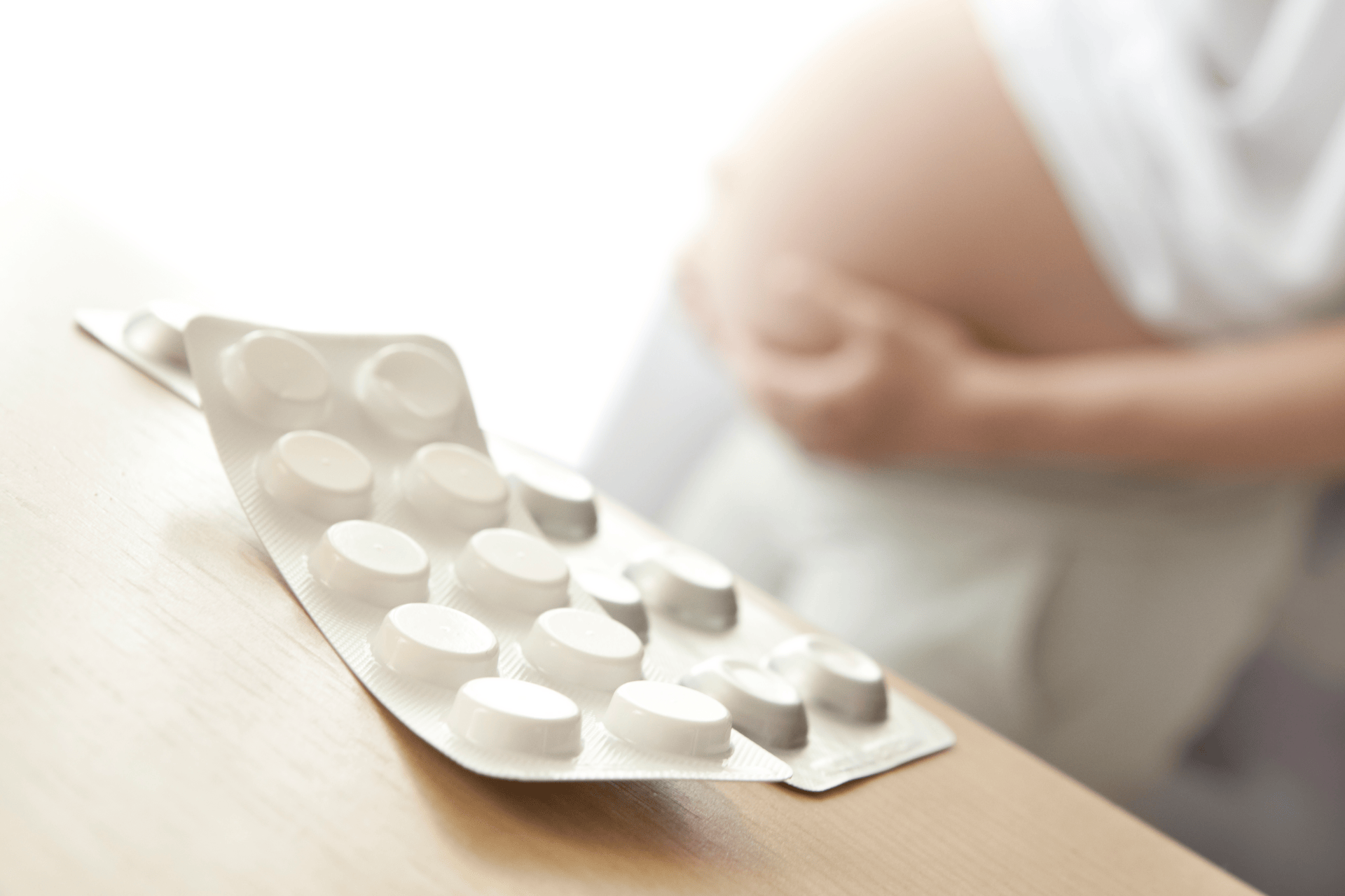 Blister pack of white tablets resting on a wooden surface, with a pregnant woman in the background.