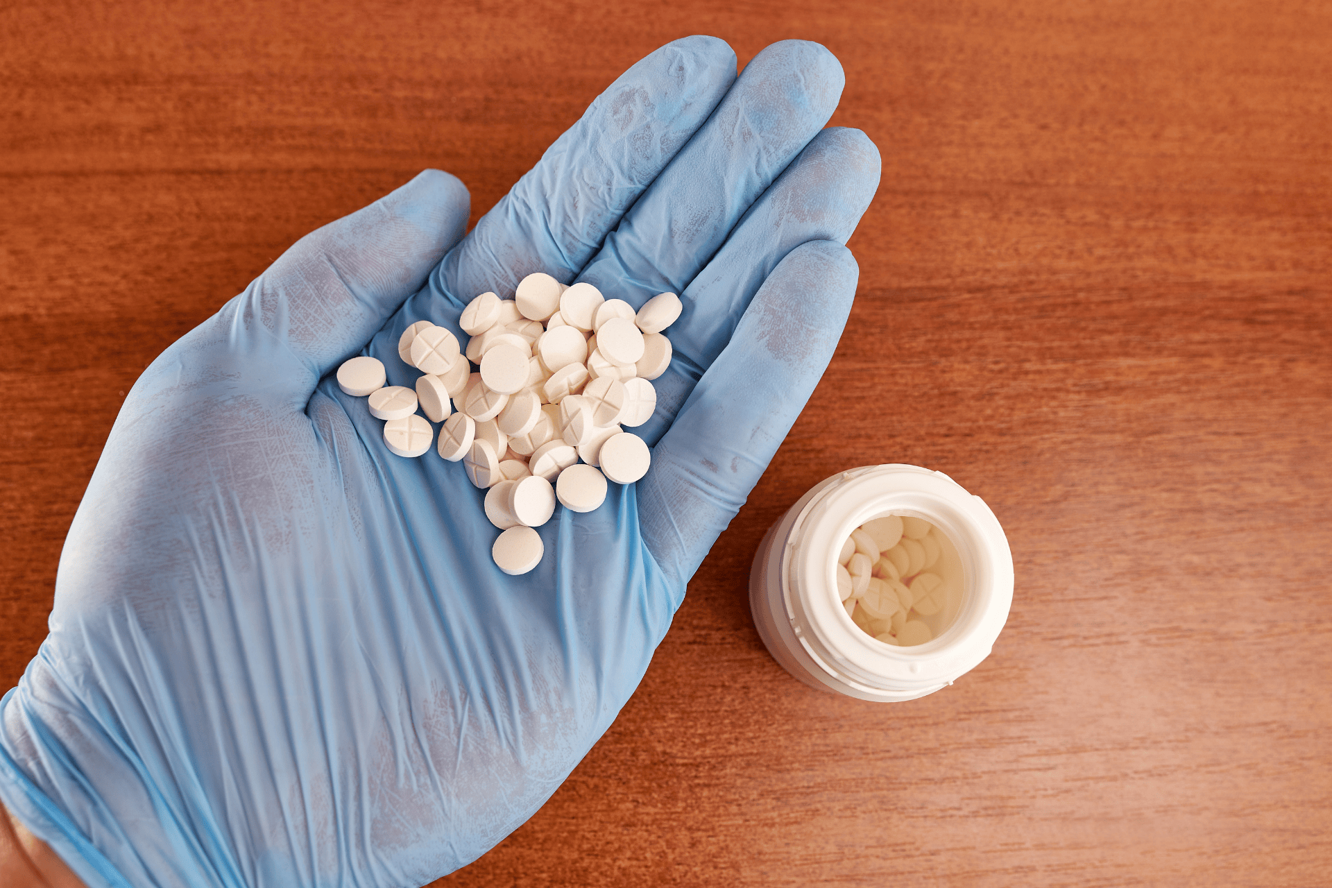 A gloved hand holding many small white tablets next to an open pill bottle on a wooden surface.