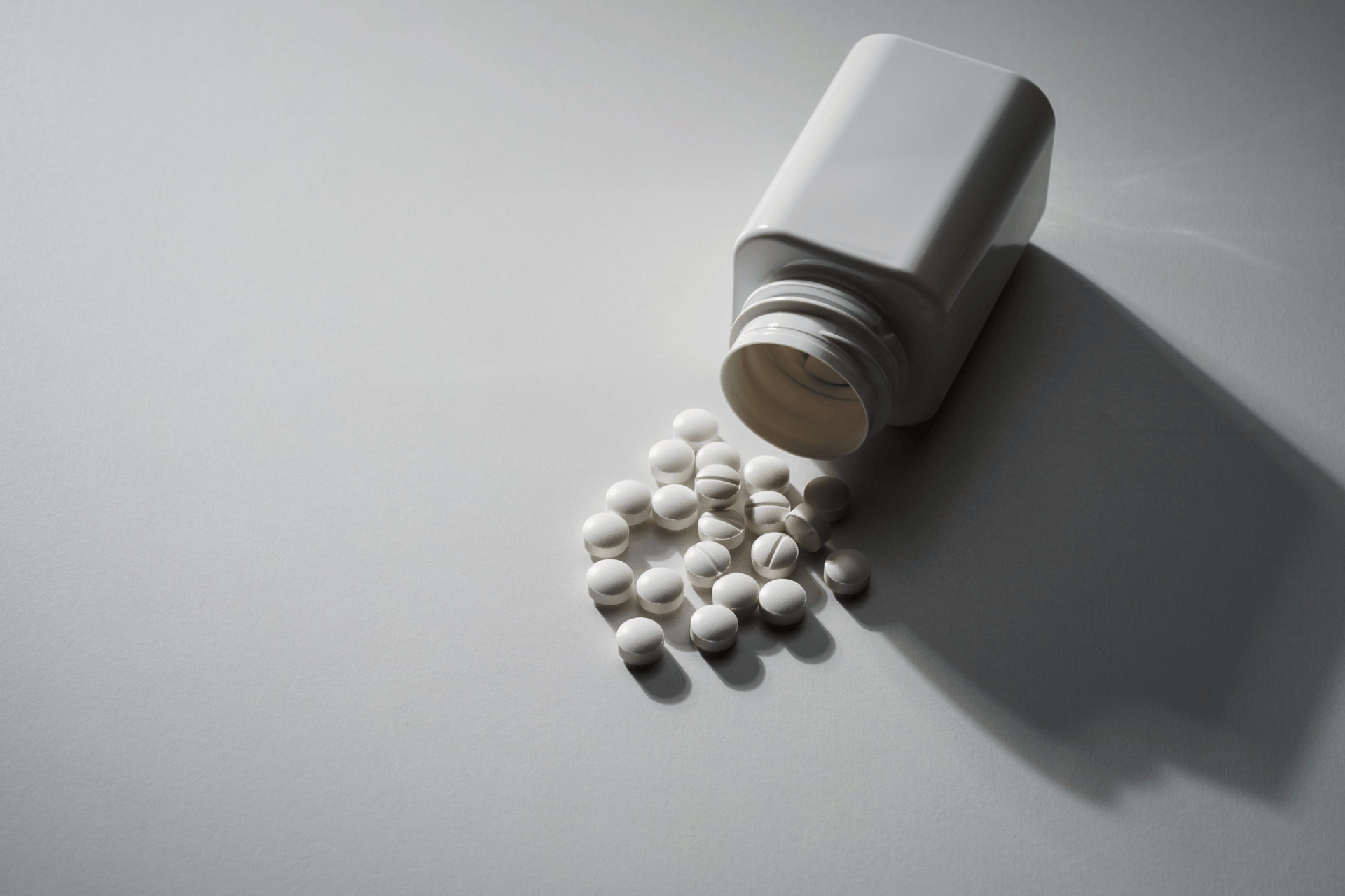 White pill bottle tipped over, spilling round tablets onto a gray surface.