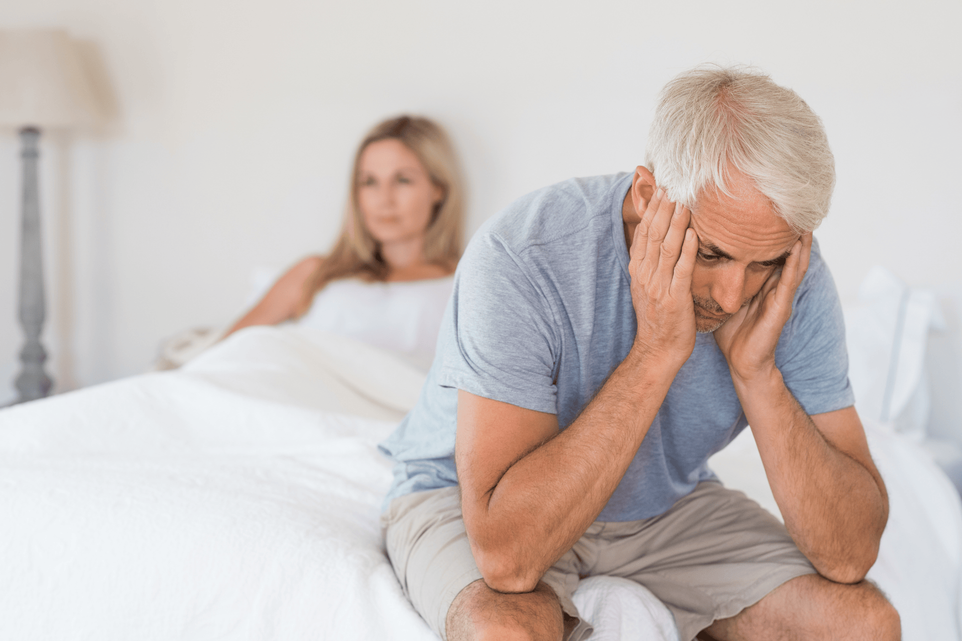 Older man sitting on a bed holding his head, partner blurred in the background.