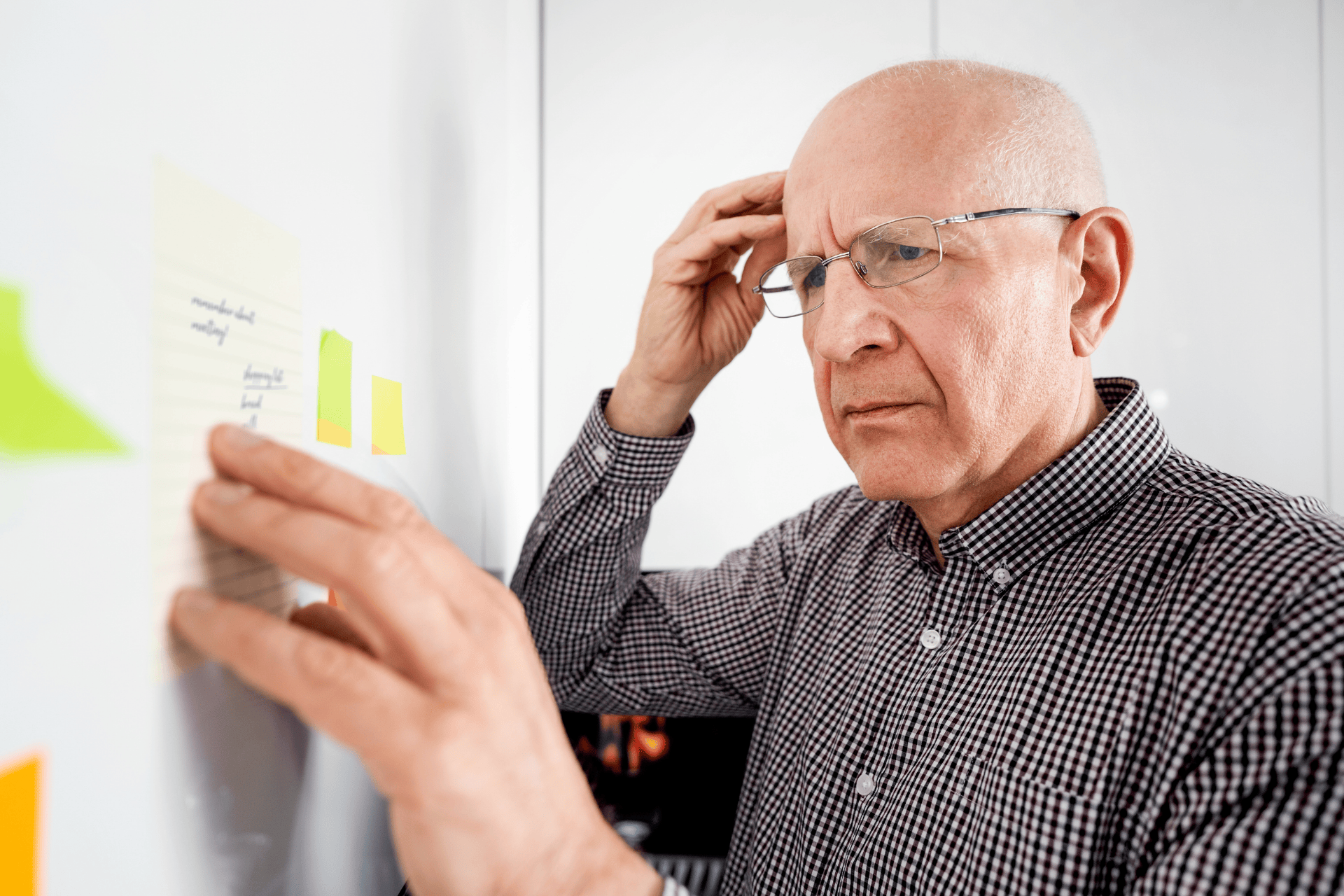 An older man wearing glasses looks closely at notes on a board while holding his head, appearing thoughtful or concerned.