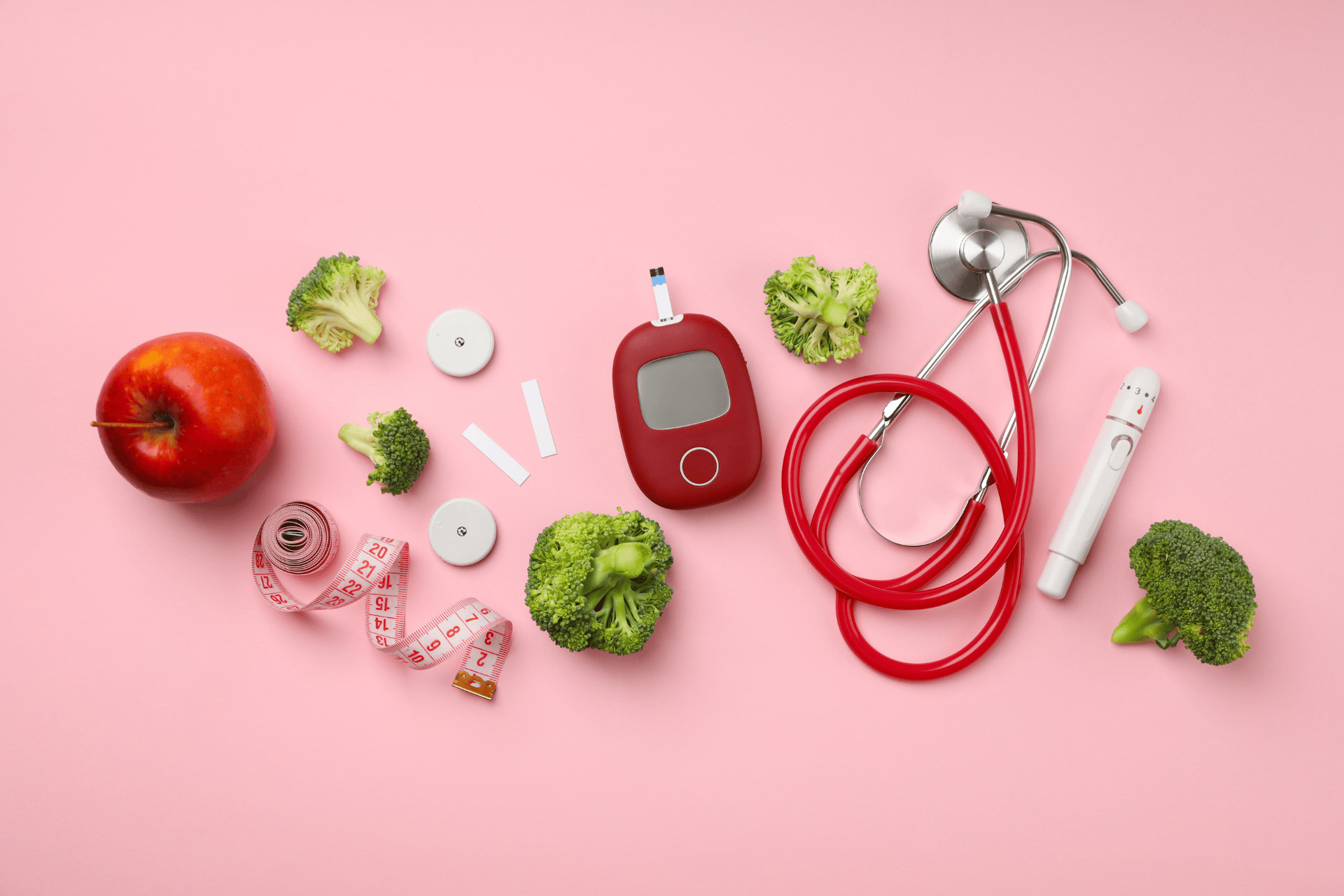 Diabetes care items arranged on a pink background, including a glucose meter, test strips, a stethoscope, an apple, broccoli, and measuring tape.