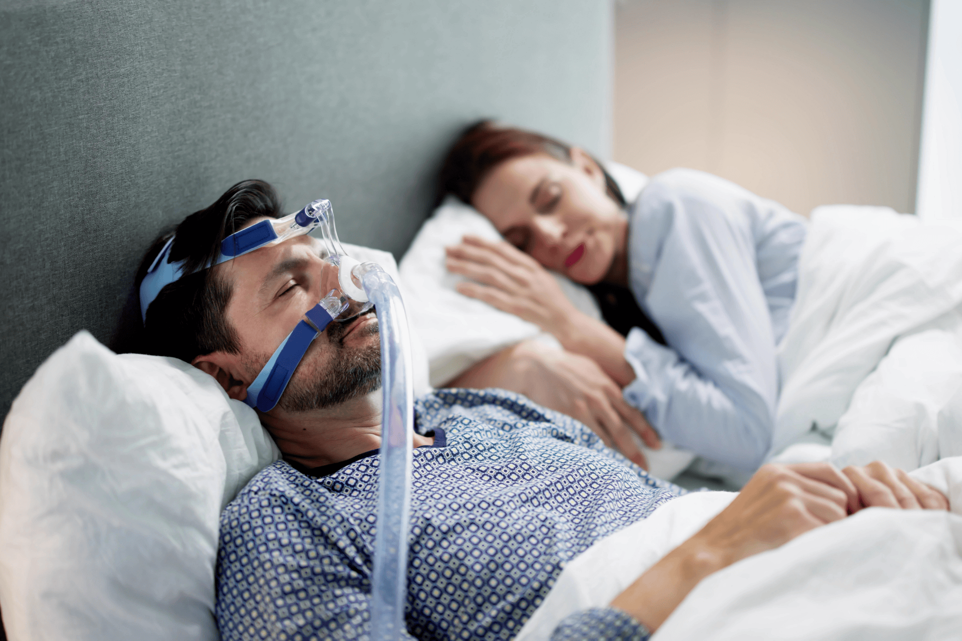 A man sleeping with a CPAP mask for sleep apnea while a partner lies beside him in bed.