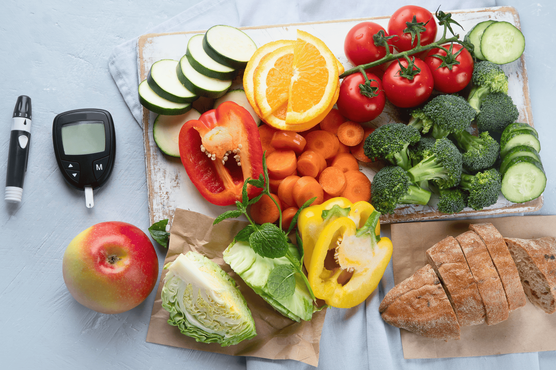 A selection of fresh vegetables, fruit, and bread alongside a glucose meter and lancing device, representing healthy eating for diabetes.