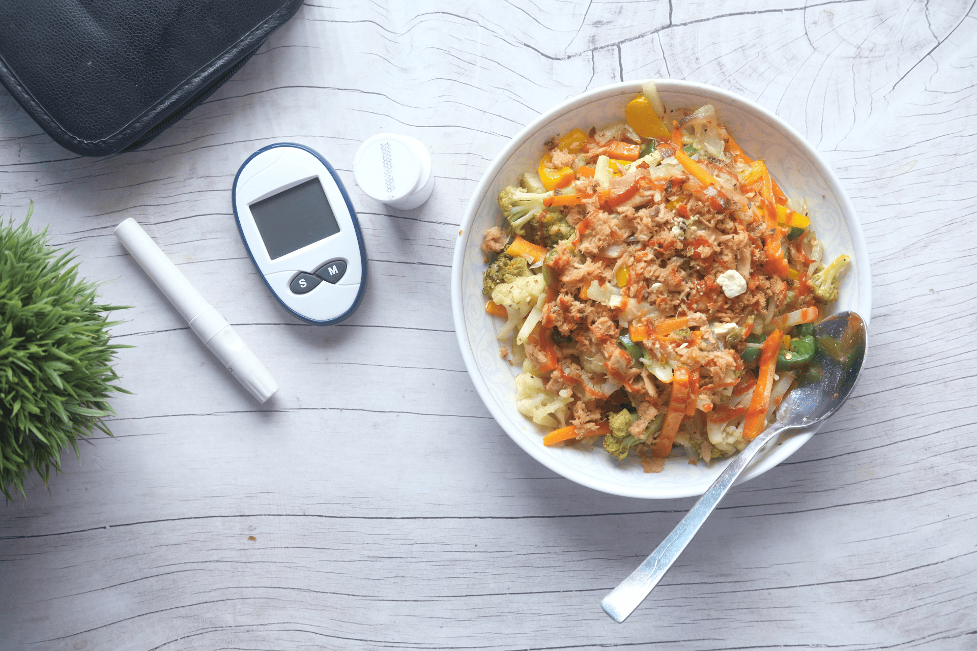 A bowl of mixed vegetables topped with shredded tuna on a light wooden table, beside a glucose meter, test strip container, lancet pen, and spoon, suggesting a diabetes-friendly meal.