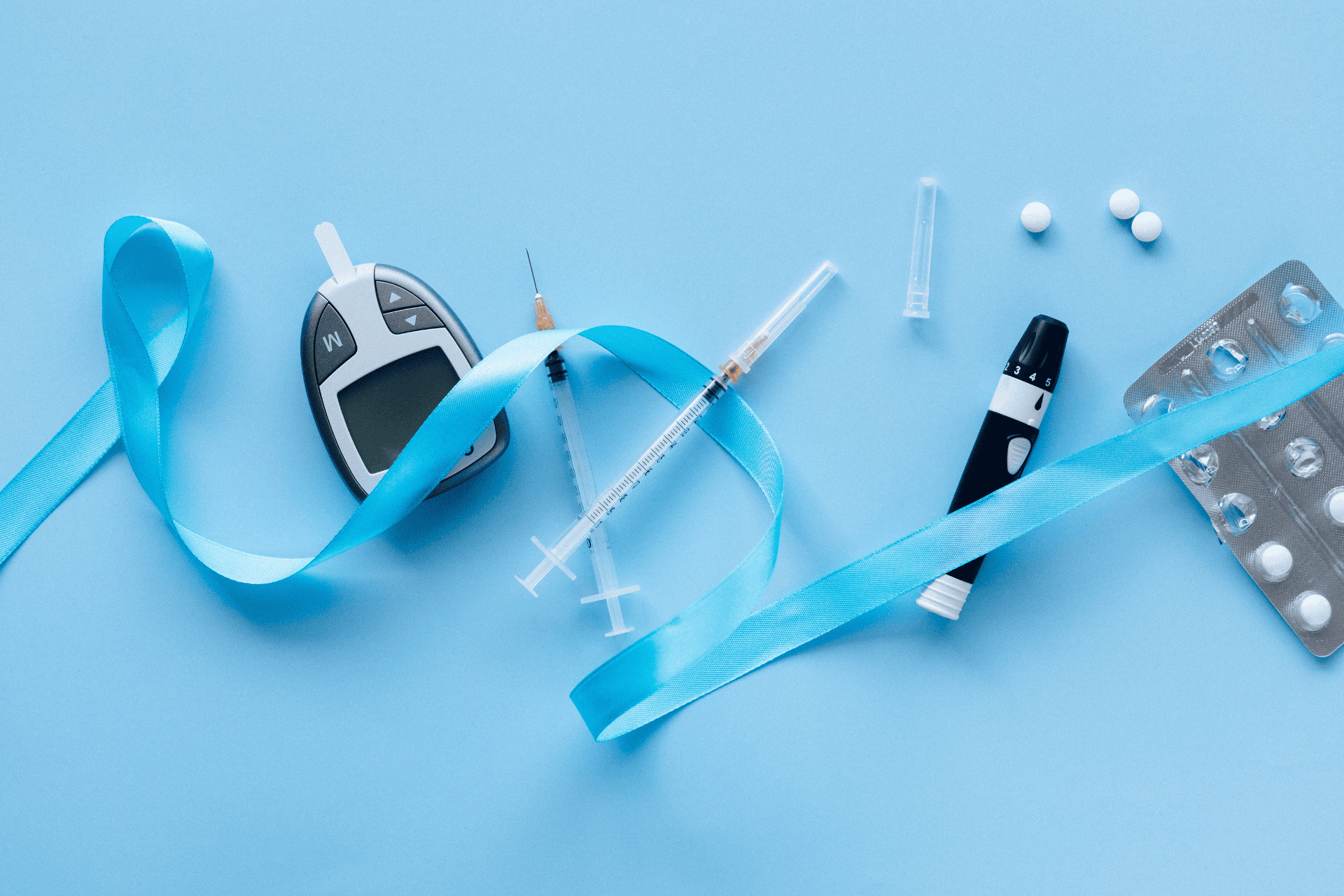 Diabetes care items on a blue background, including a glucose meter, insulin syringes, an insulin pen, test strips, pills, and a blue awareness ribbon.