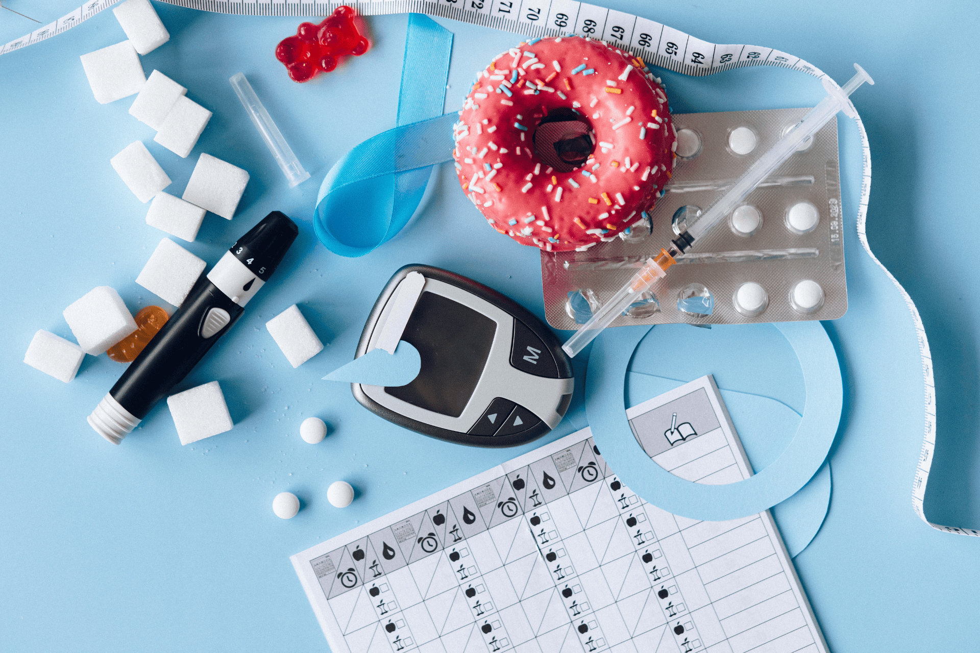Flat lay of diabetes supplies, glucose meter, insulin pen and syringe, sugar cubes, pills, and a pink donut on a blue background with a measuring tape and log sheet.