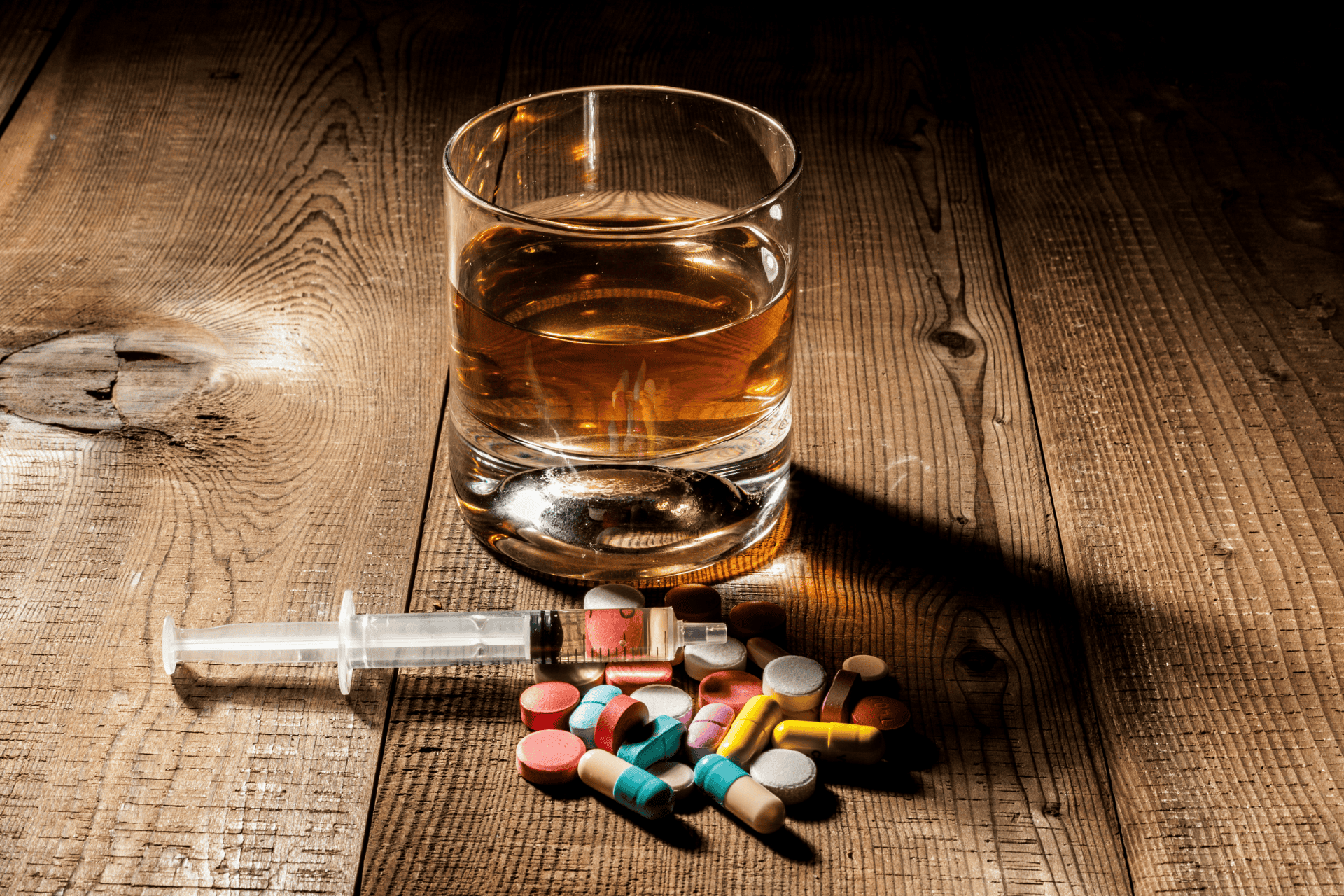 A glass of alcohol beside a syringe and assorted pills on a wooden surface, symbolizing substance misuse, addiction, and the dangerous interaction between alcohol and injectable or oral drugs.