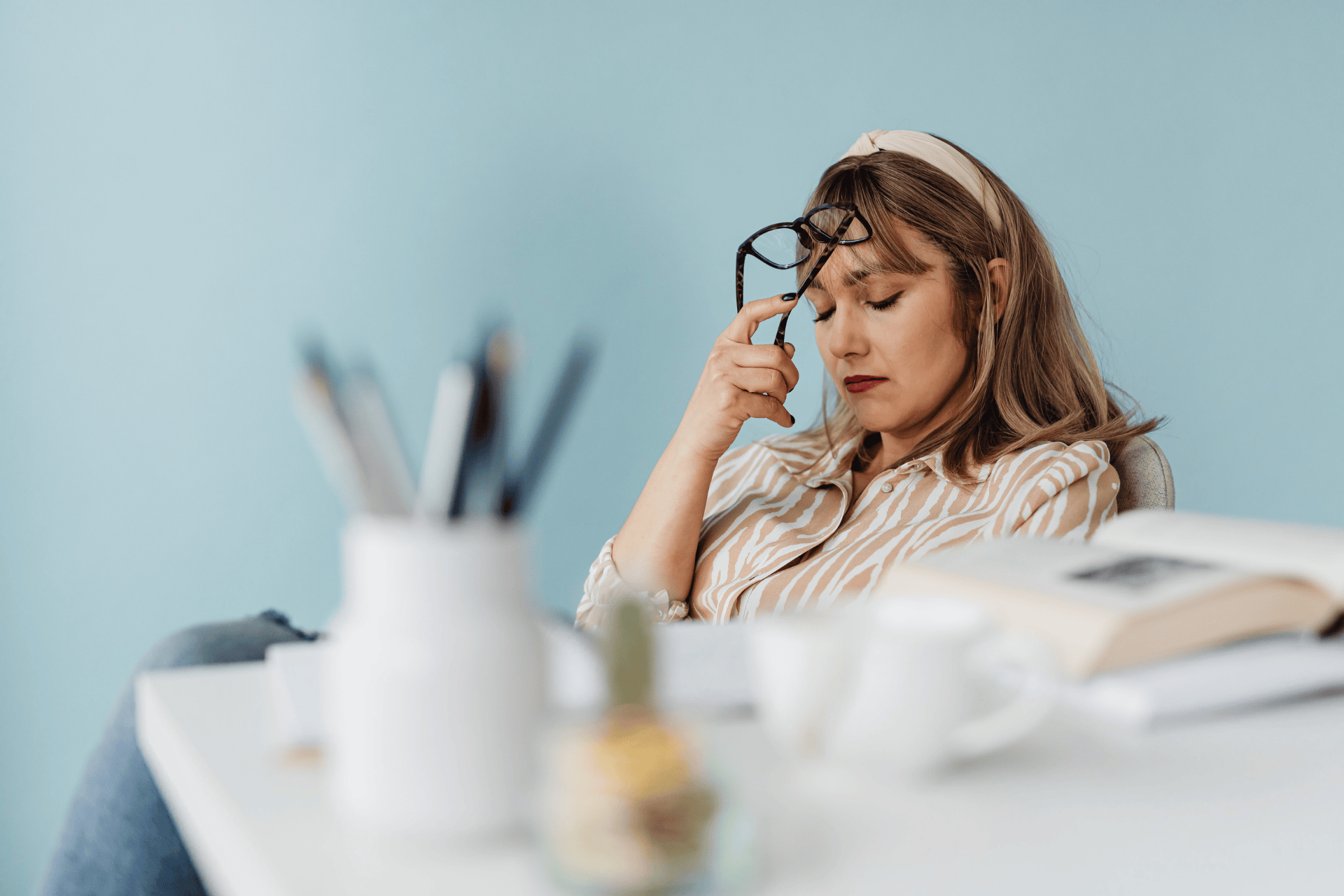 Woman sitting at a desk, eyes closed, holding her glasses and appearing stressed or tired.