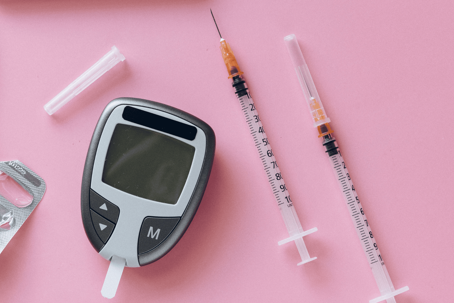 Blood glucose meter and insulin syringes on a pink background.