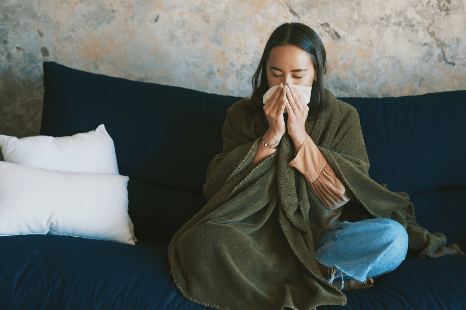 Woman wrapped in a blanket sitting on a couch, blowing her nose with a tissue during flu recovery.
