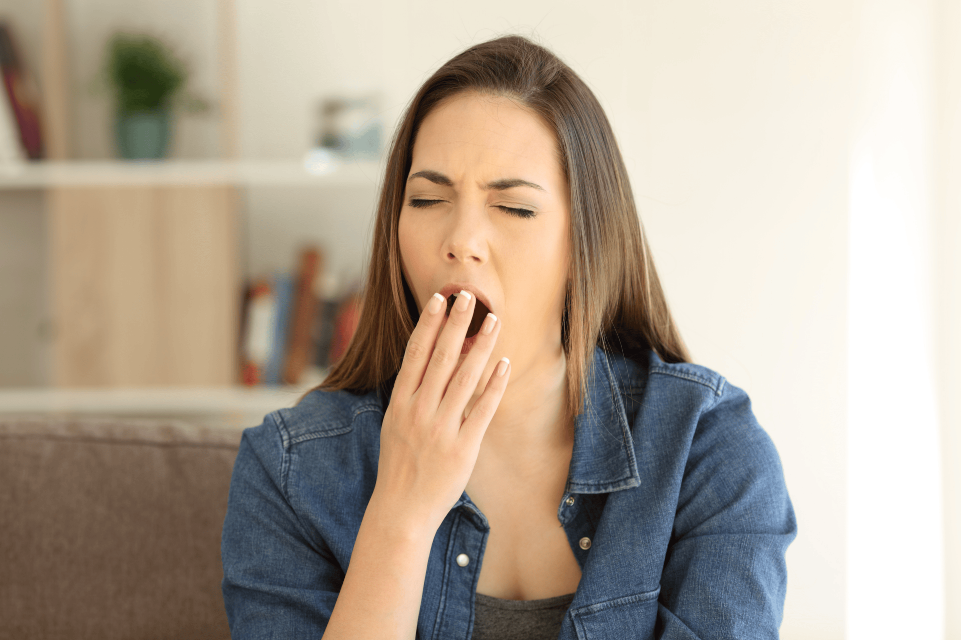 Woman sitting indoors, yawning with her hand over her mouth, showing fatigue.