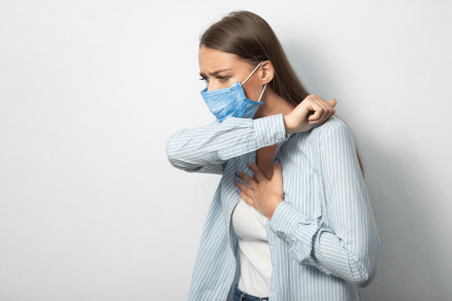 Woman wearing a face mask coughing into her elbow, showing COVID-related cough symptoms.