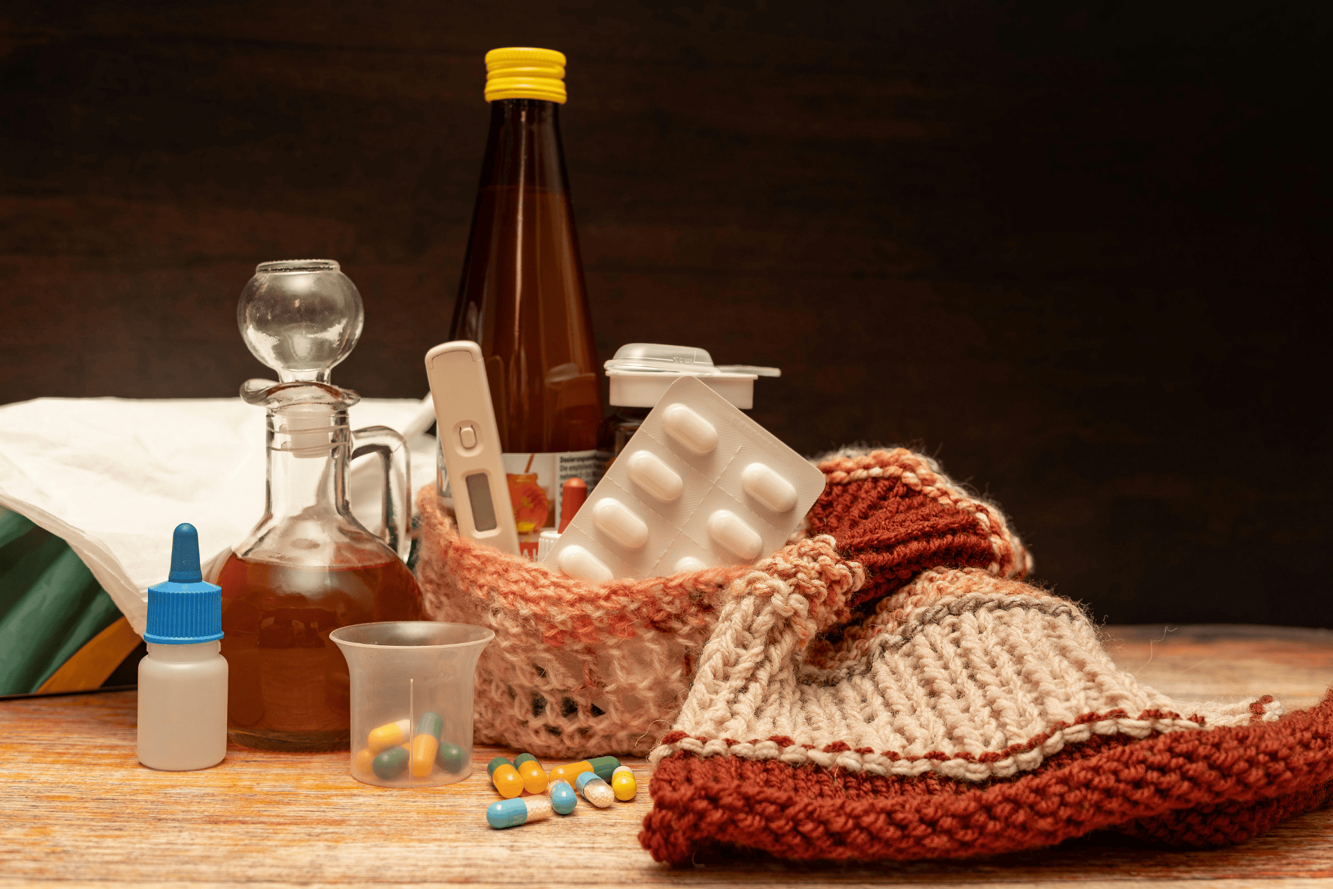 Flu medicine and home remedies on a table, including pills, cough syrup, a thermometer, nasal drops, and a knitted scarf.
