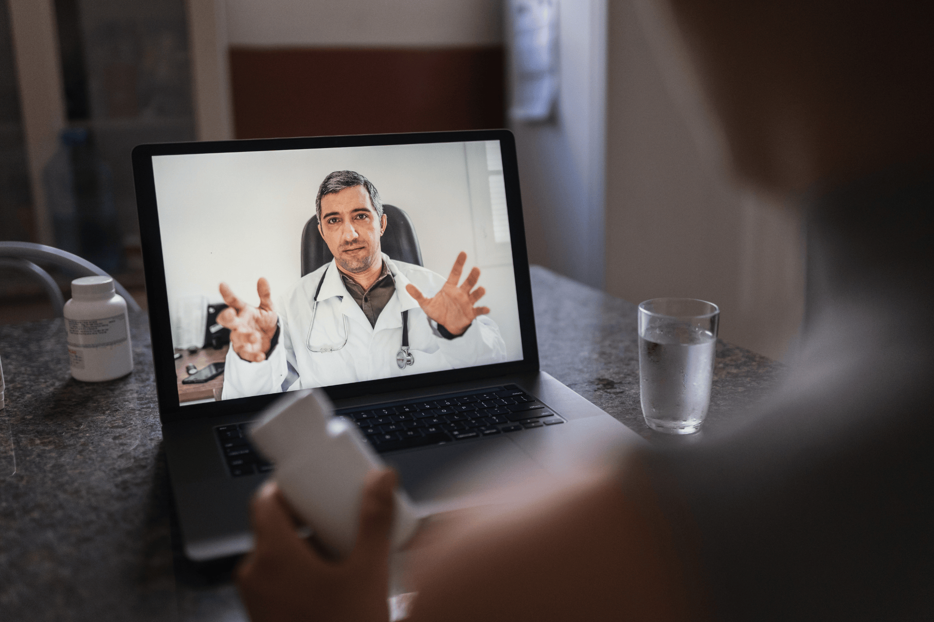 Person holding a medication bottle while having an online video consultation with a doctor displayed on a laptop screen.