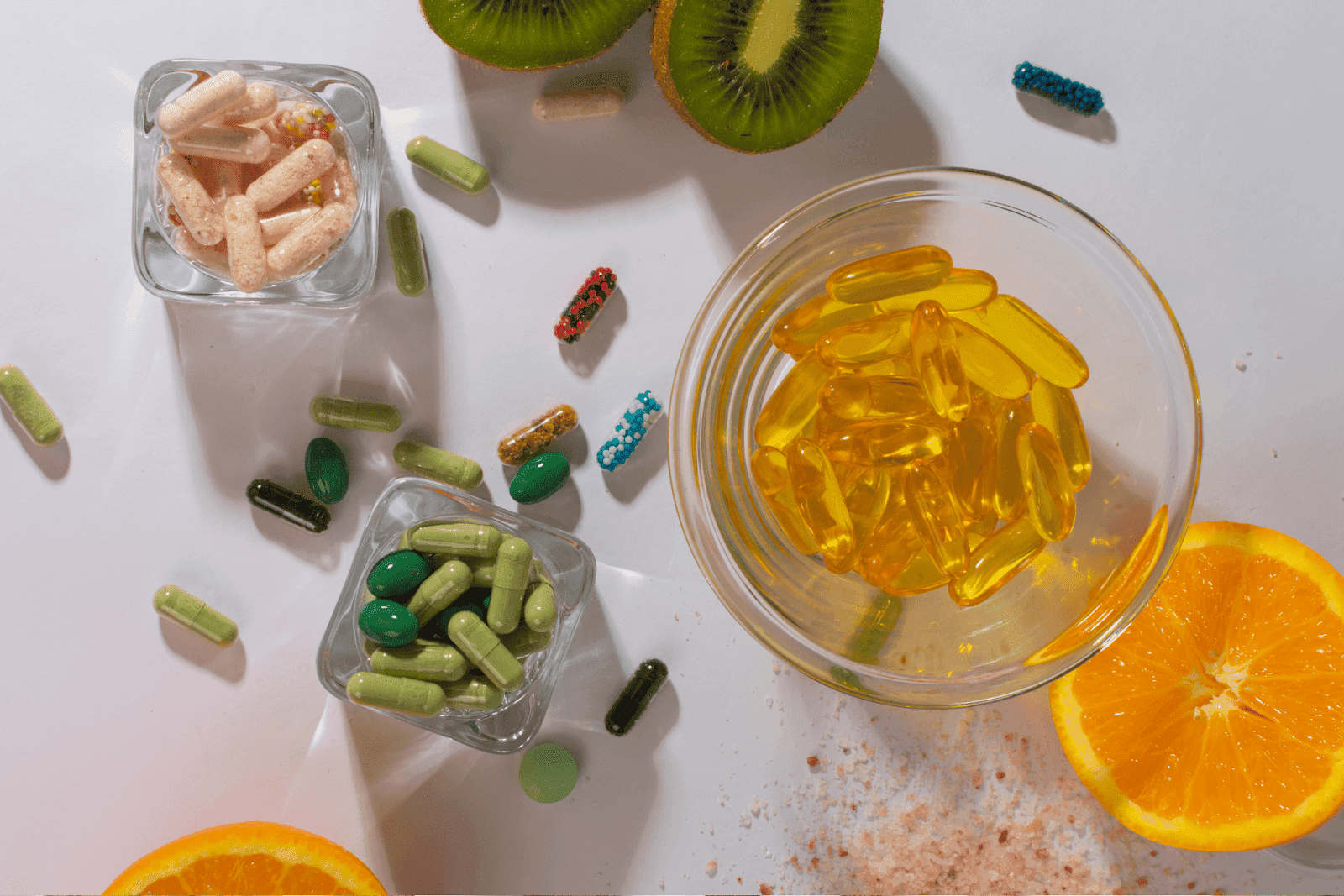Variety of colorful vitamin capsules and pills scattered with slices of orange and kiwi on a white surface.