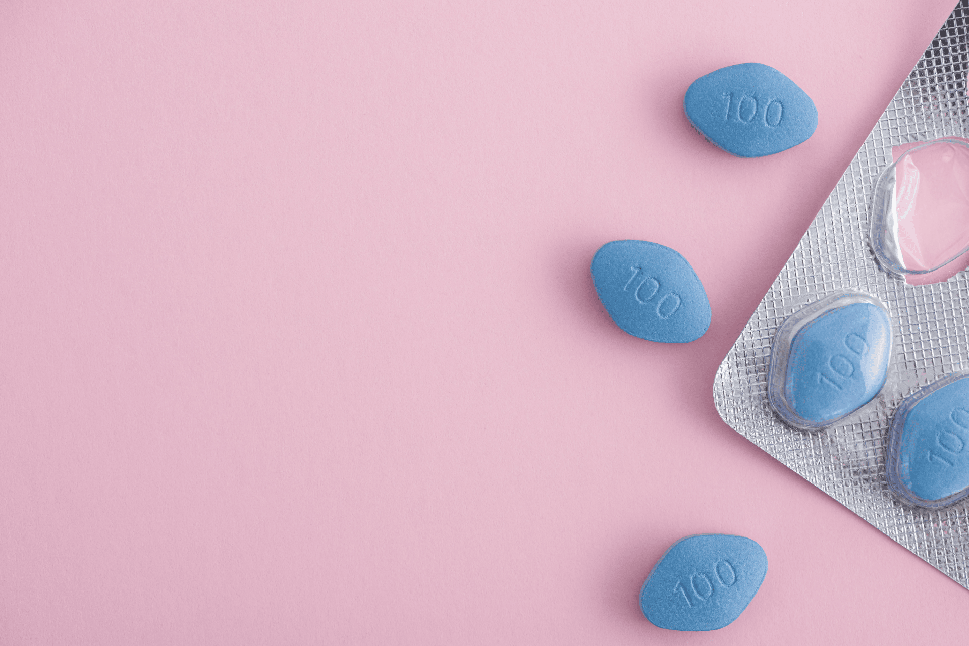Blue Viagra pills scattered next to a blister pack on a pink background.