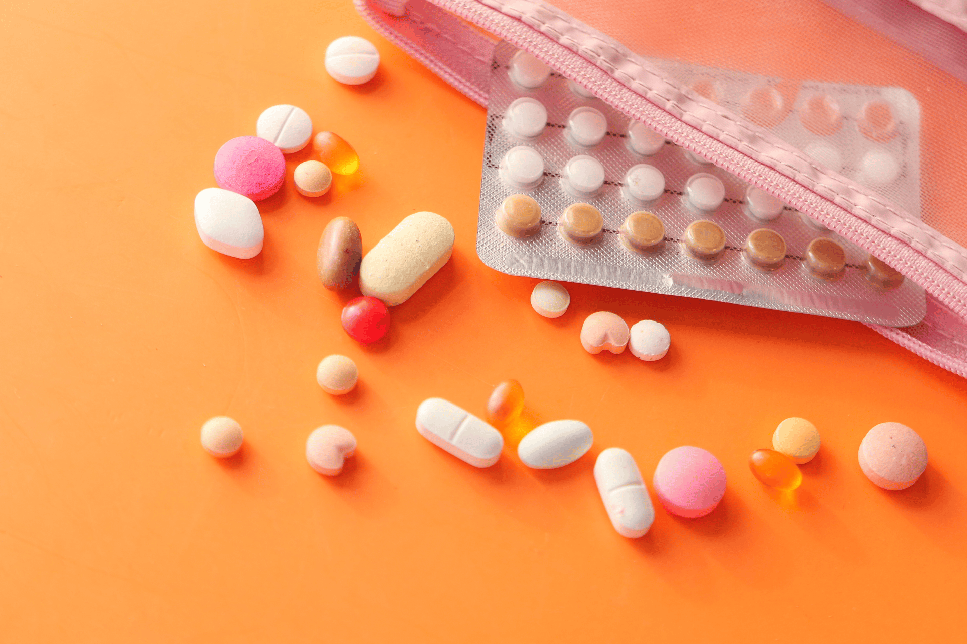 Various colorful pills and a blister pack of tablets spilling out of a pink pouch onto an orange surface.