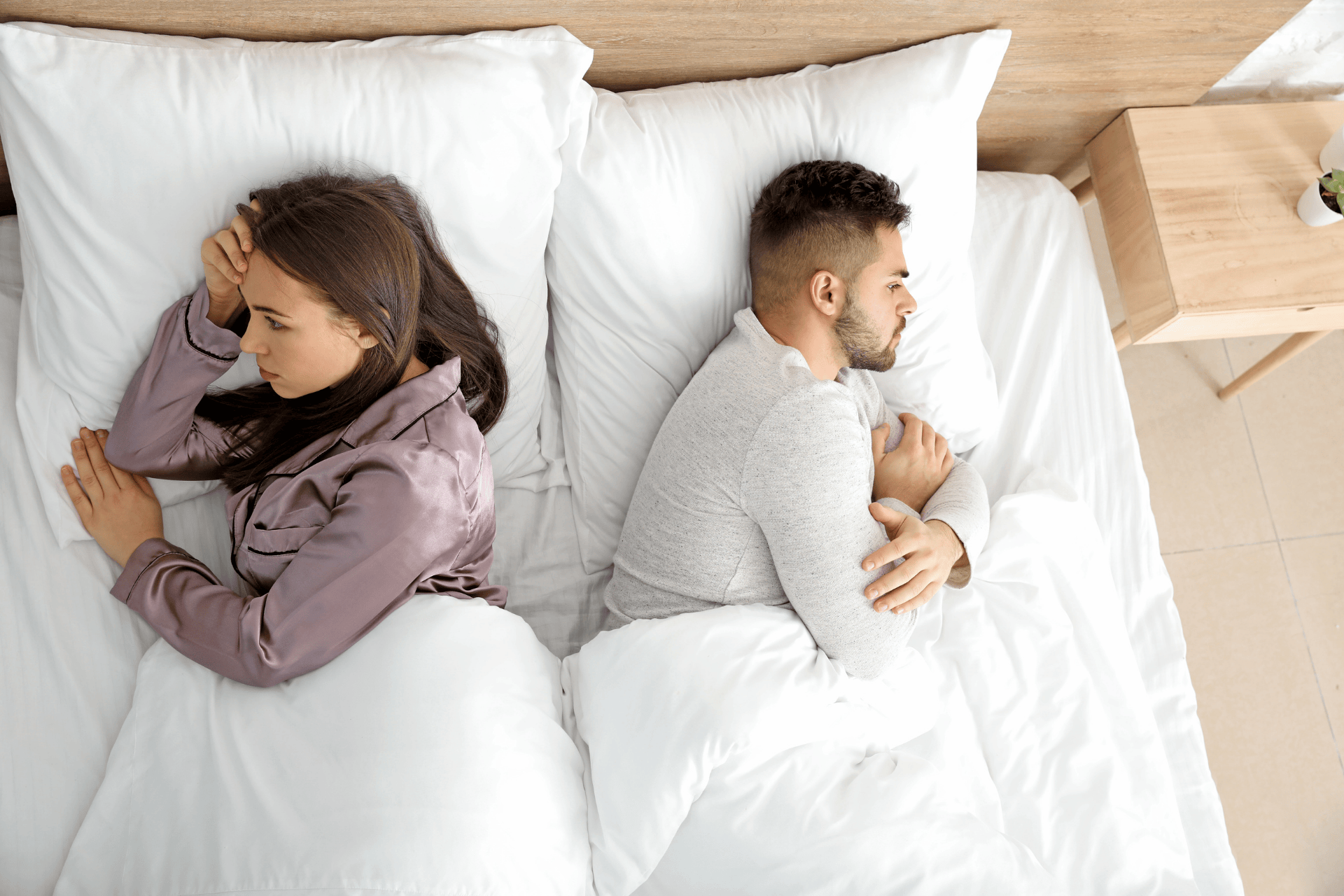 Couple lying in bed facing away from each other, looking upset and distant.
