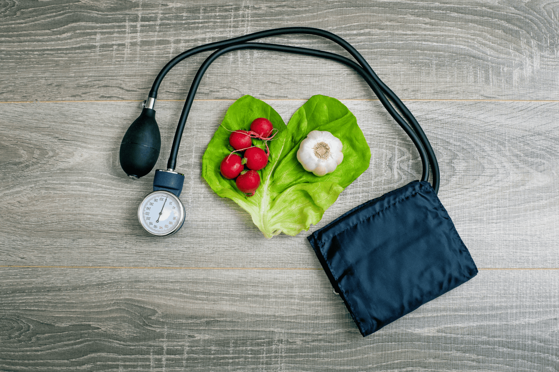 Blood pressure monitor next to lettuce leaves shaped like a heart, radishes, and garlic on a wooden surface.