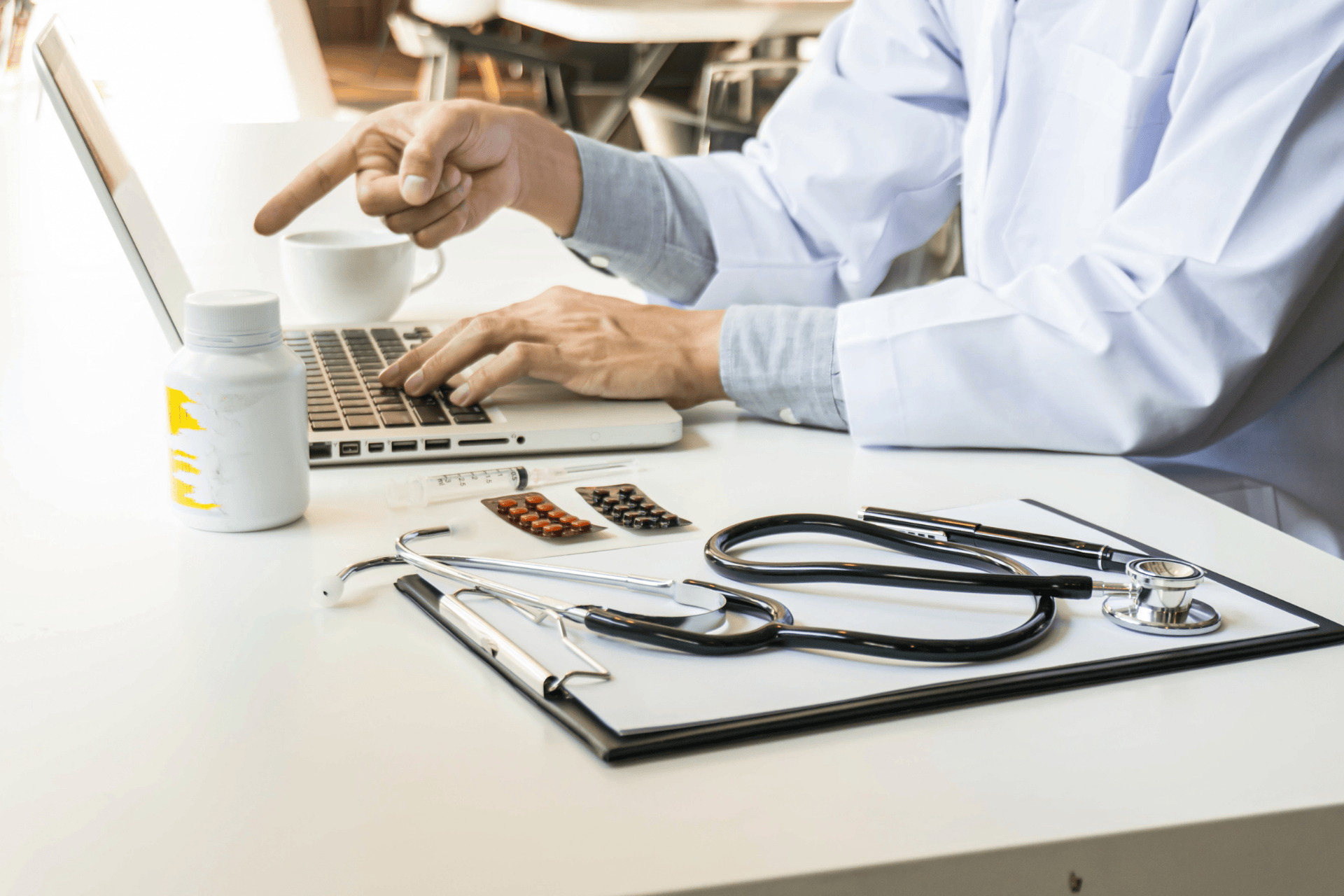 Doctor using a laptop with medical tools, pills, and a stethoscope on the desk.