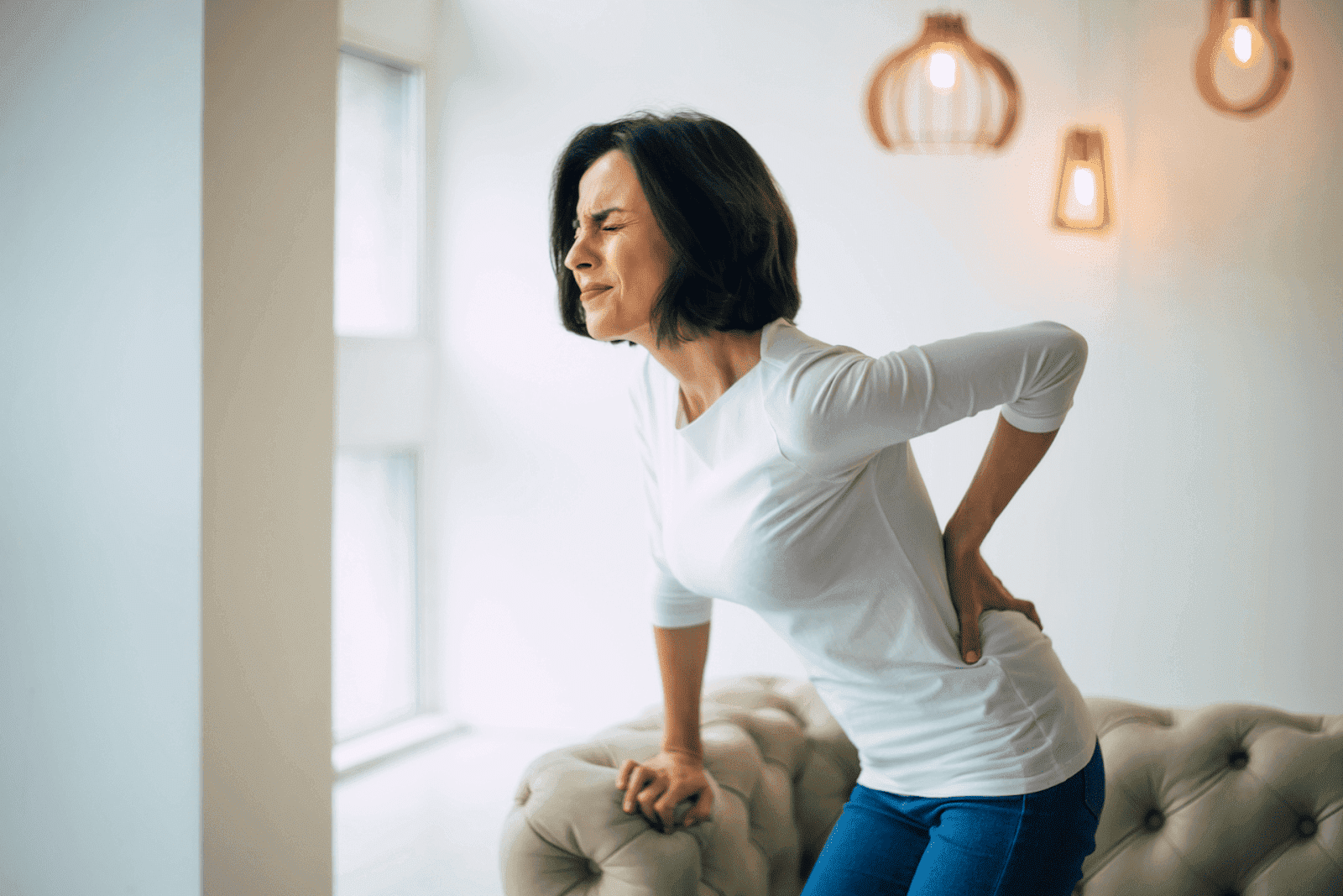 Woman grimacing in pain while holding her lower back and leaning on a couch