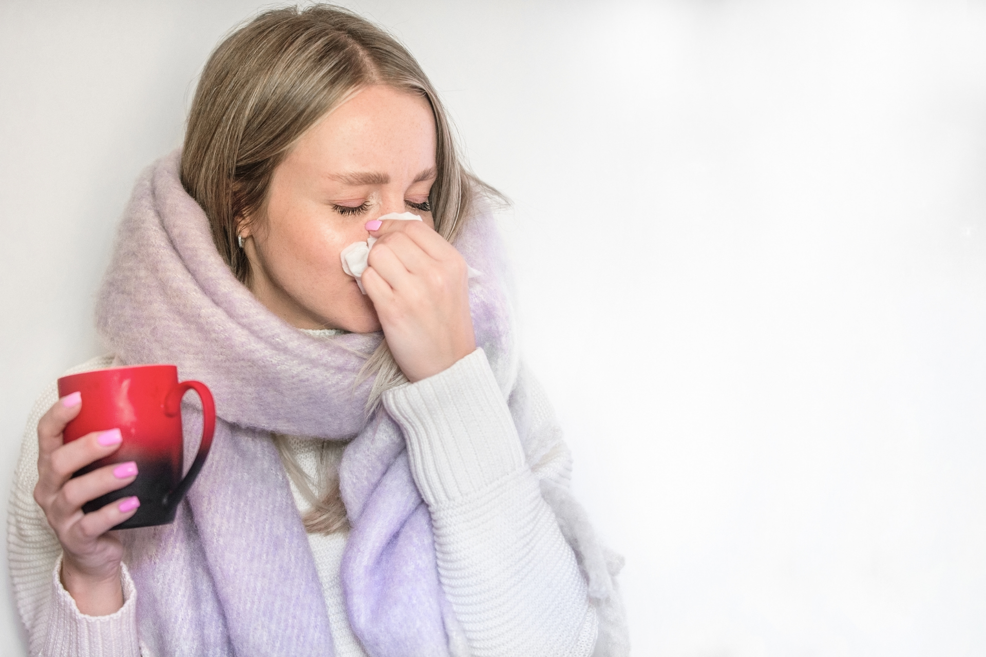 Woman holding a mug while blowing her nose, appearing to have a cold