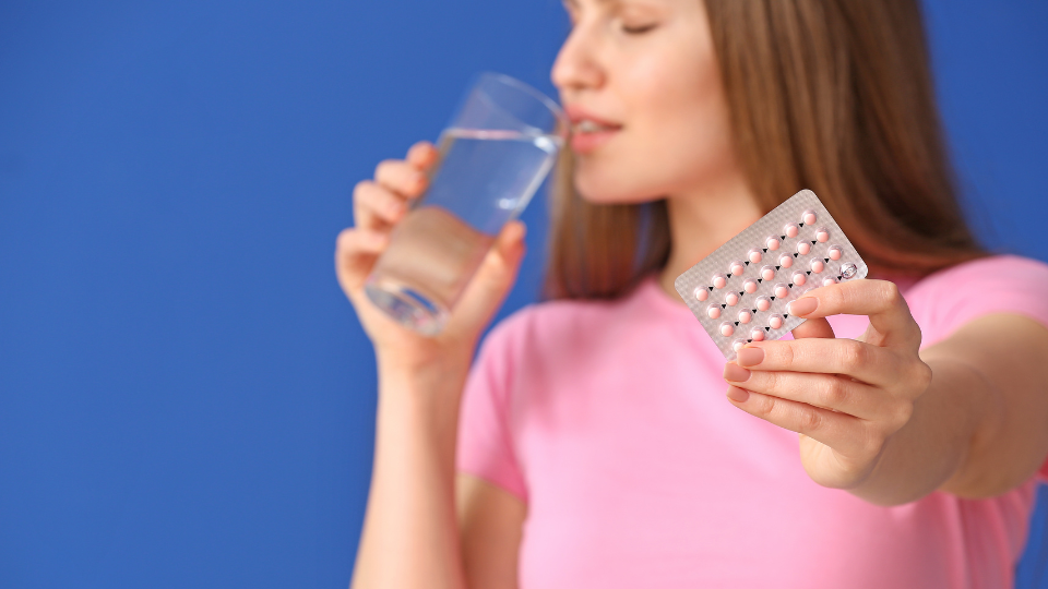 Woman drinking glass of water and holding up birth control pills.