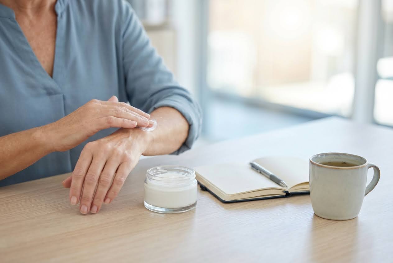 Woman in a blue shirt applying white cream to her forearm, with an open notebook and a mug on a light wooden table.