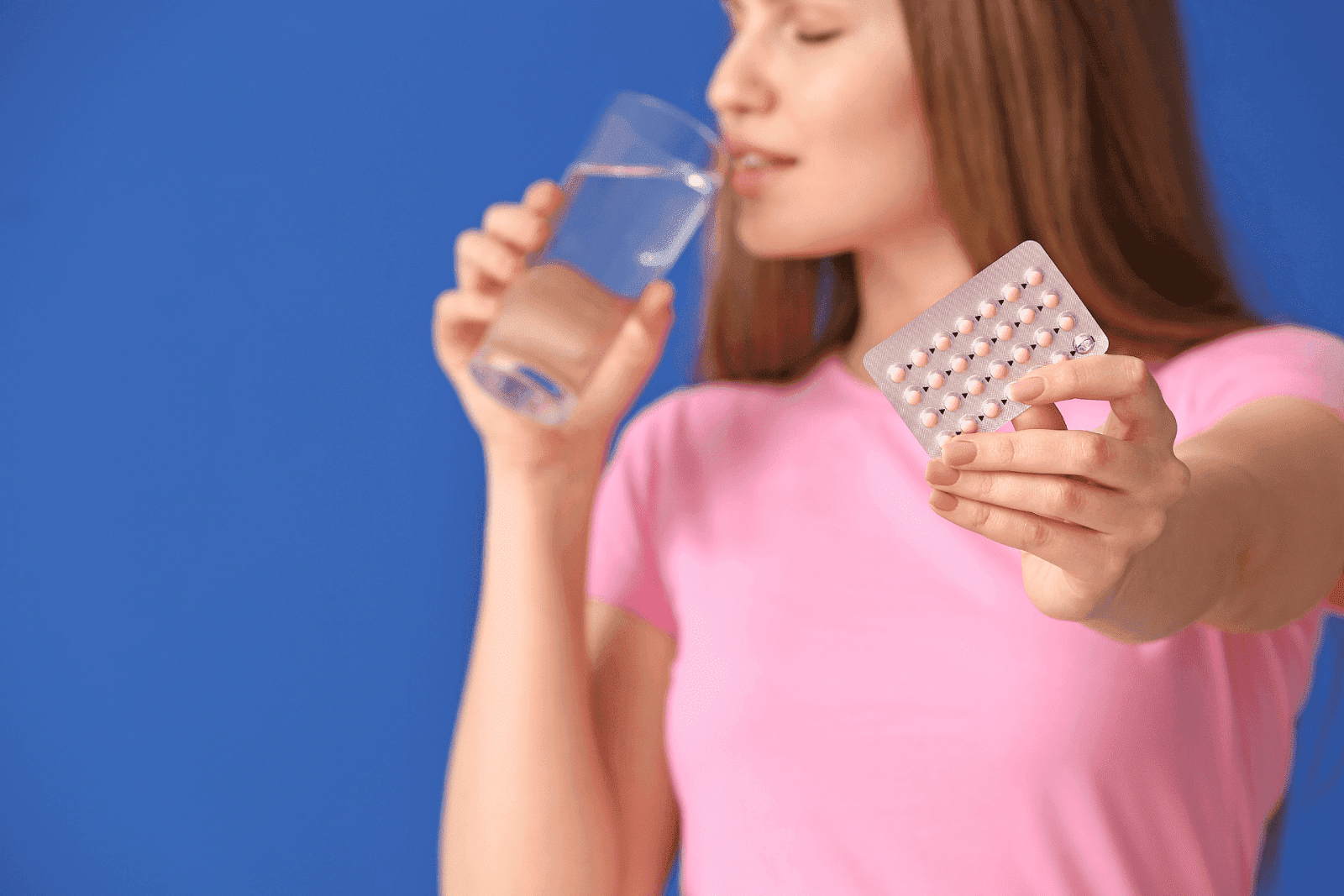 Woman in a pink shirt holding a pack of birth control pills while drinking a glass of water